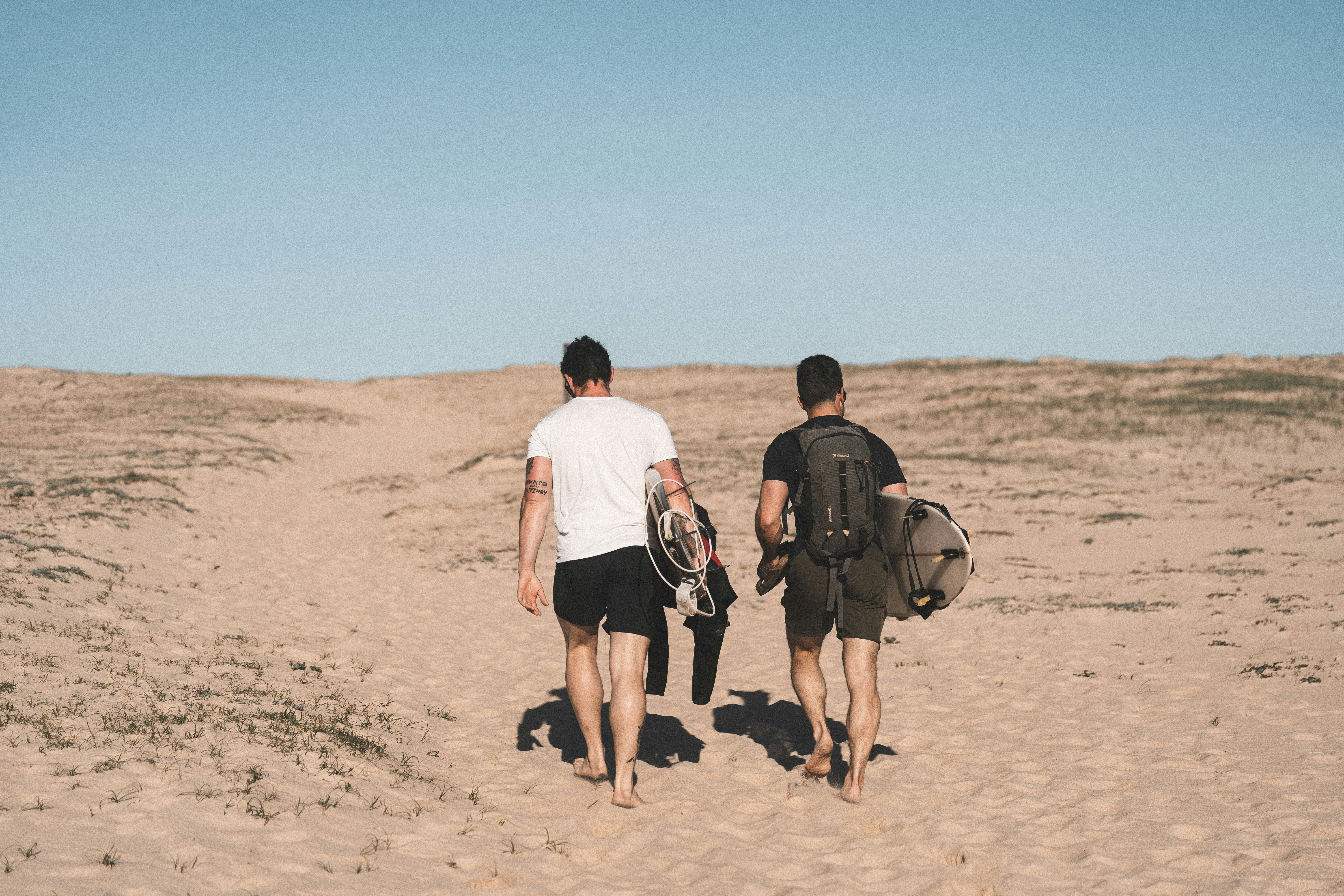 Two men walking barefoot across sand dunes carrying surfboards and a wetsuit