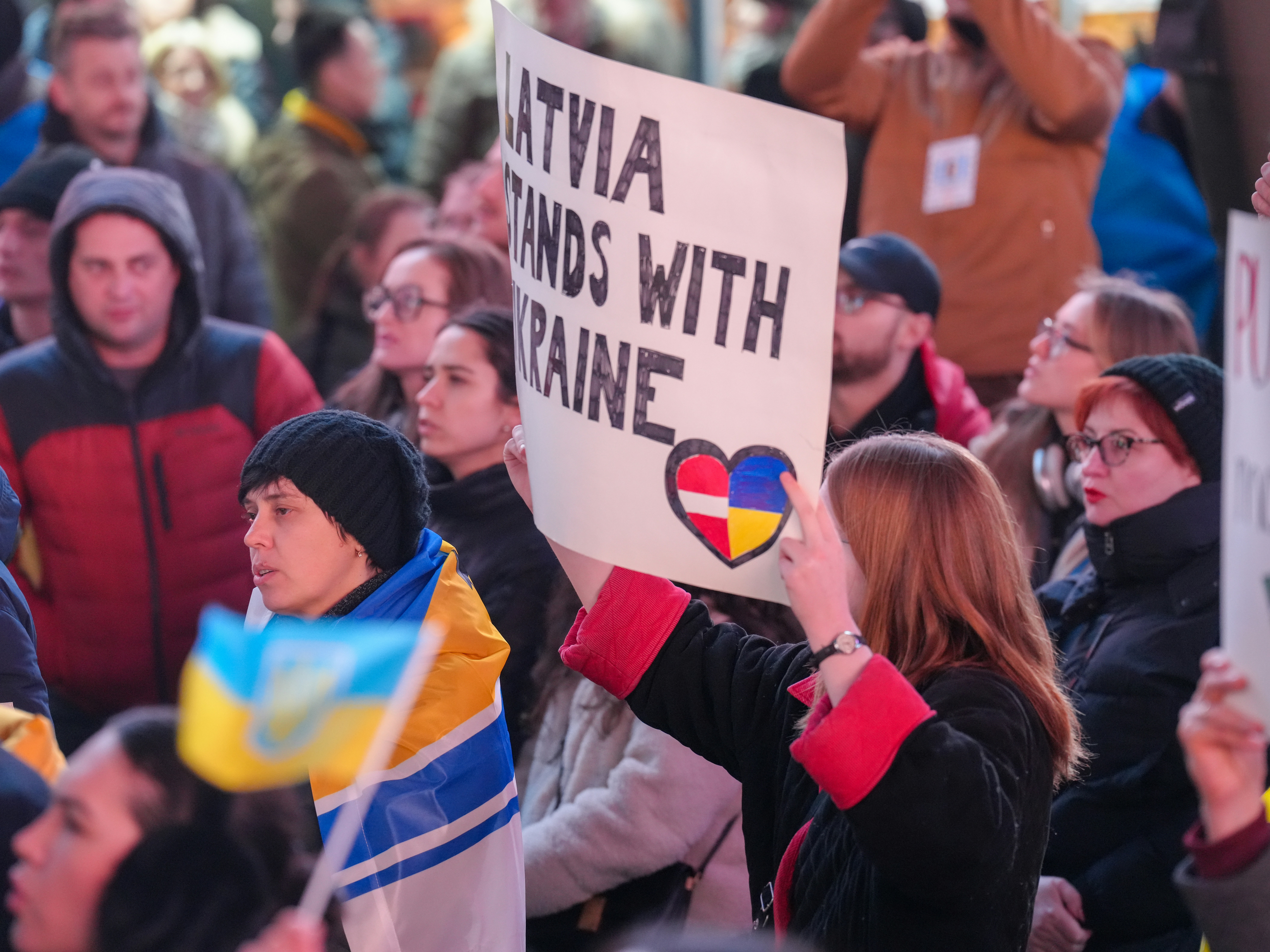 A person holding a sign reading Latvia stands with Ukraine in a crowd