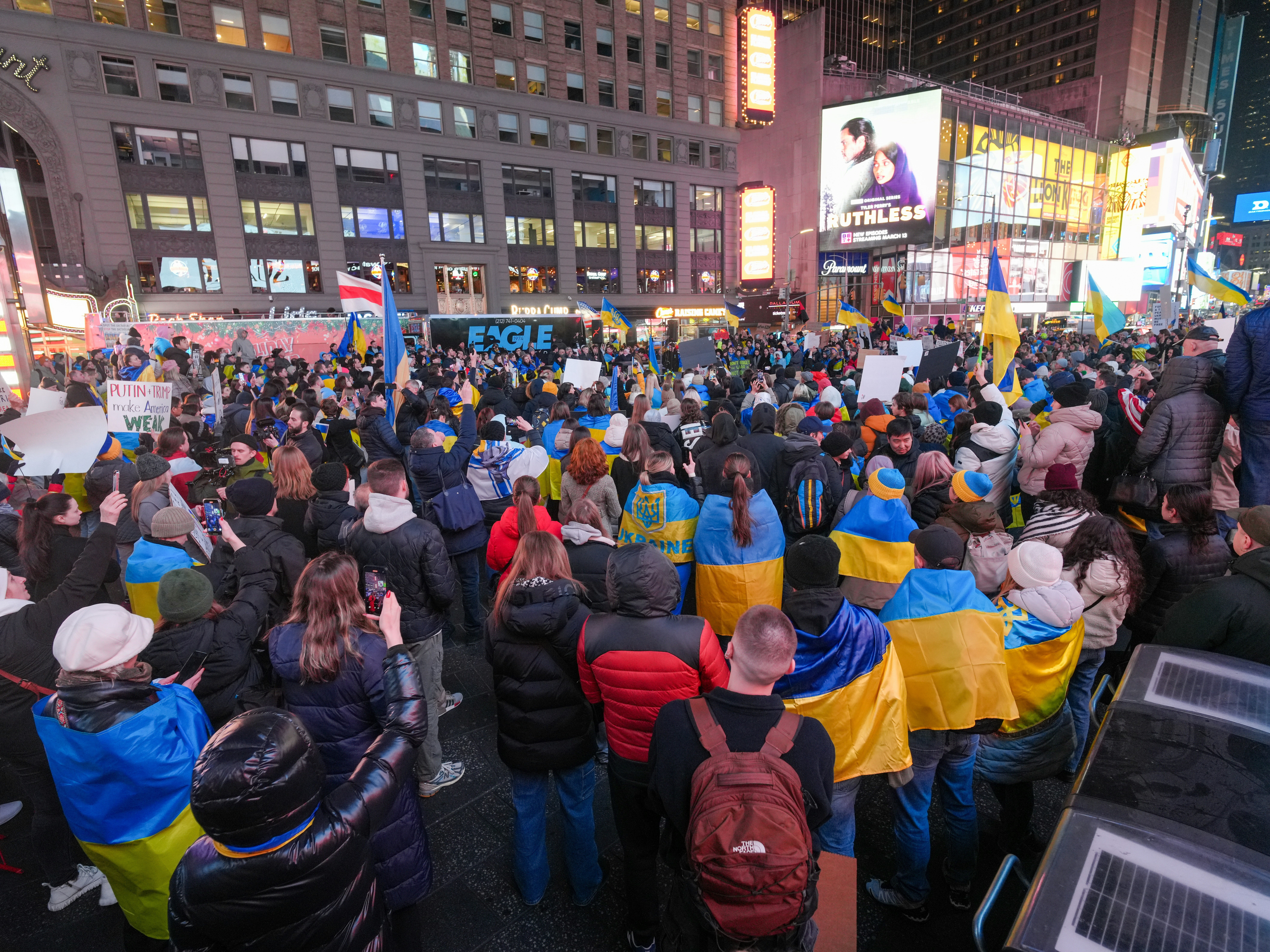 A wide view of a large rally in Times Square after dark, with protesters wrapped in Ukrainian flags, waving banners and 