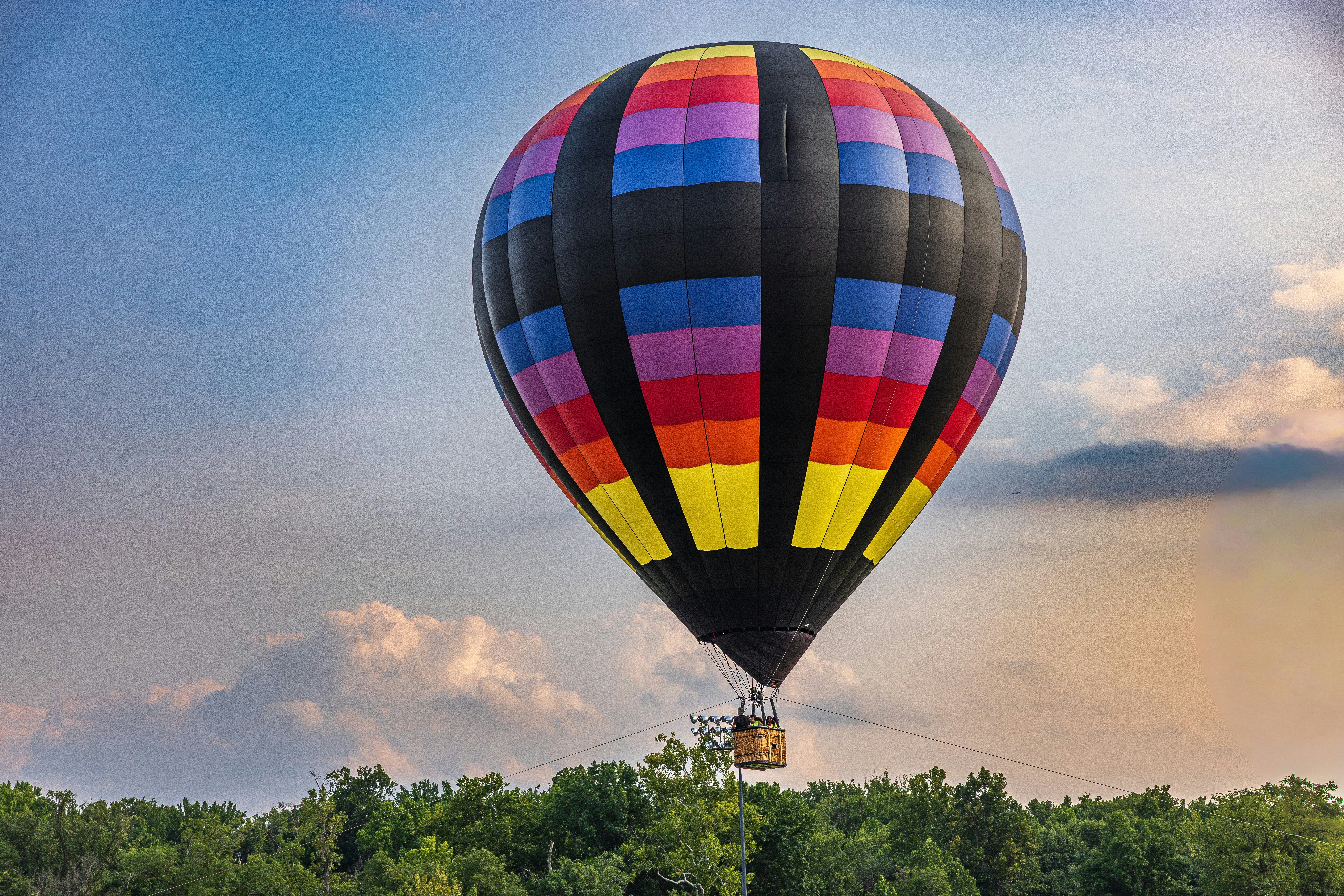 A colorful hot air balloon with black panels floating above a green forest