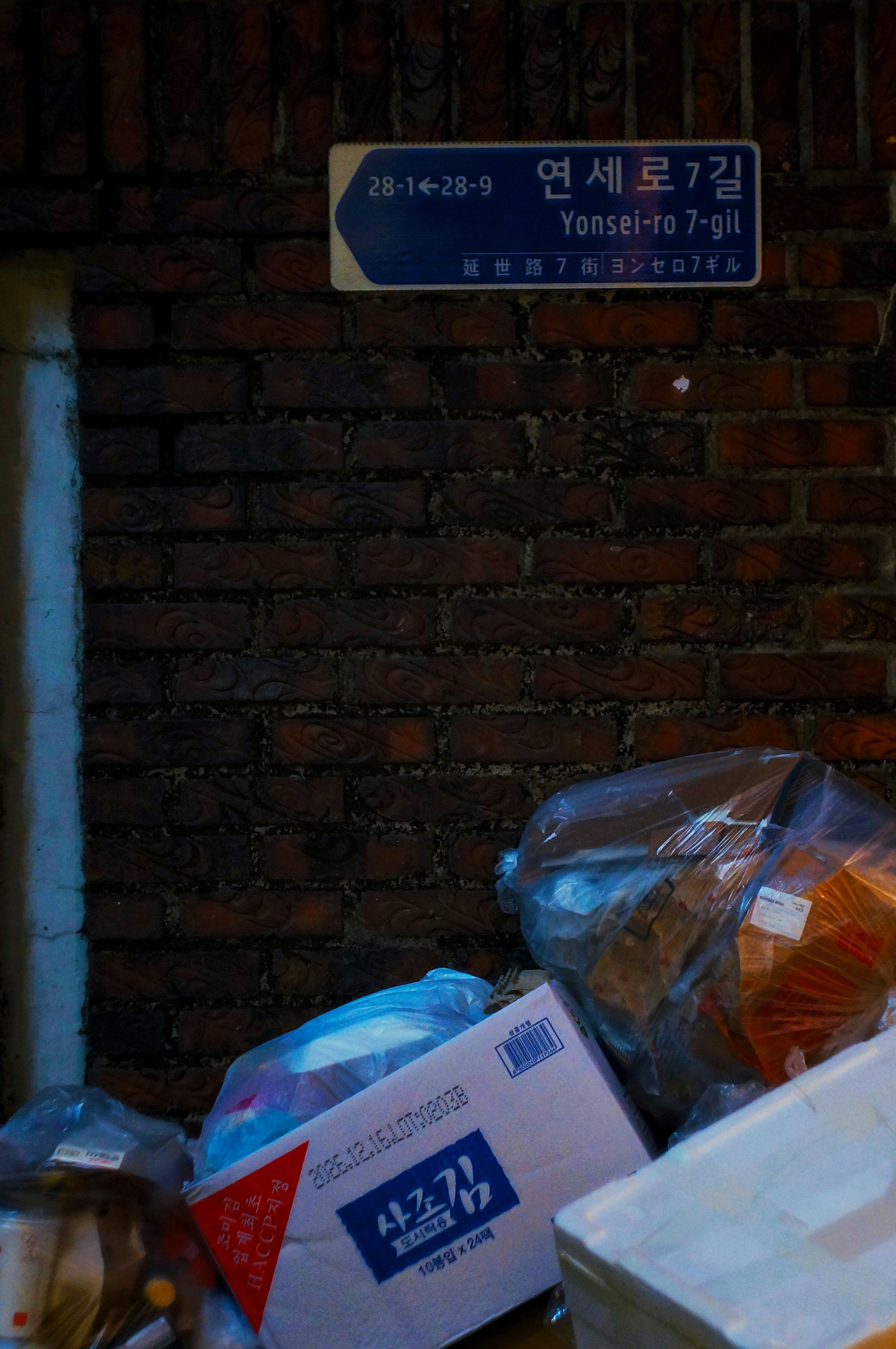 A street sign for Yonsei-ro 7-gil above a brick wall with trash bags