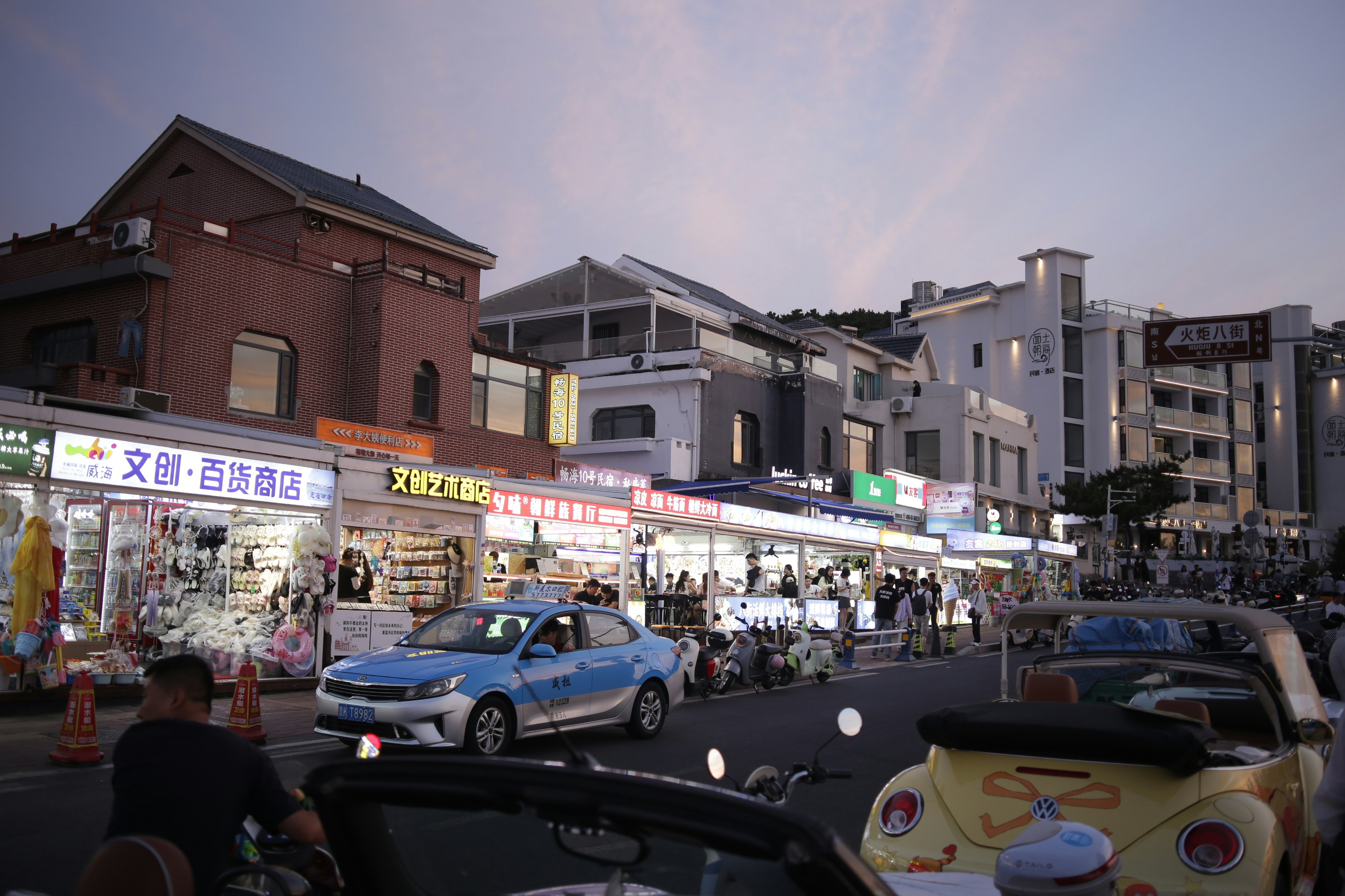 A blue taxi driving past brightly lit storefronts on a street in Weihai