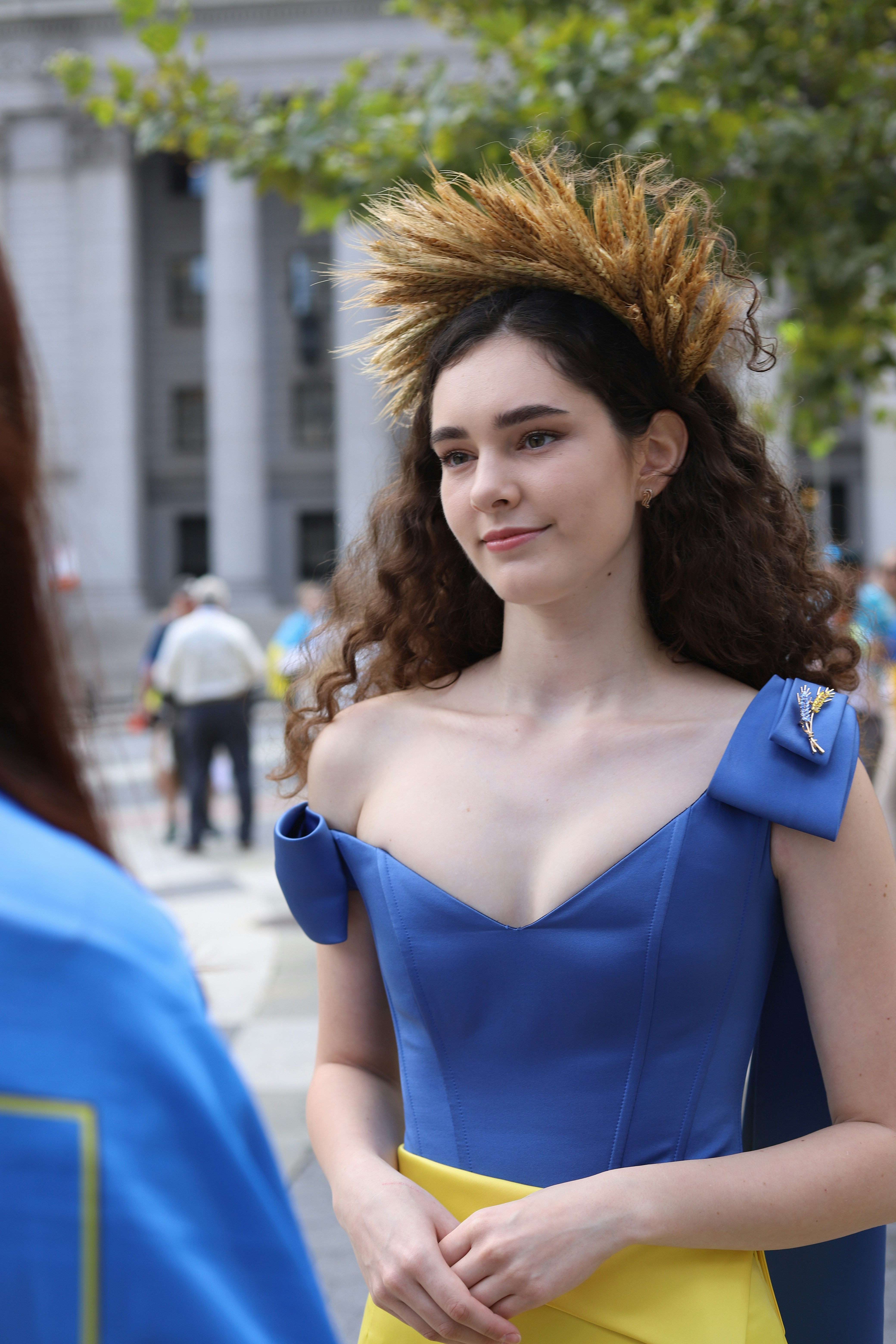 A woman with a wheat crown wearing a blue dress with a yellow sash