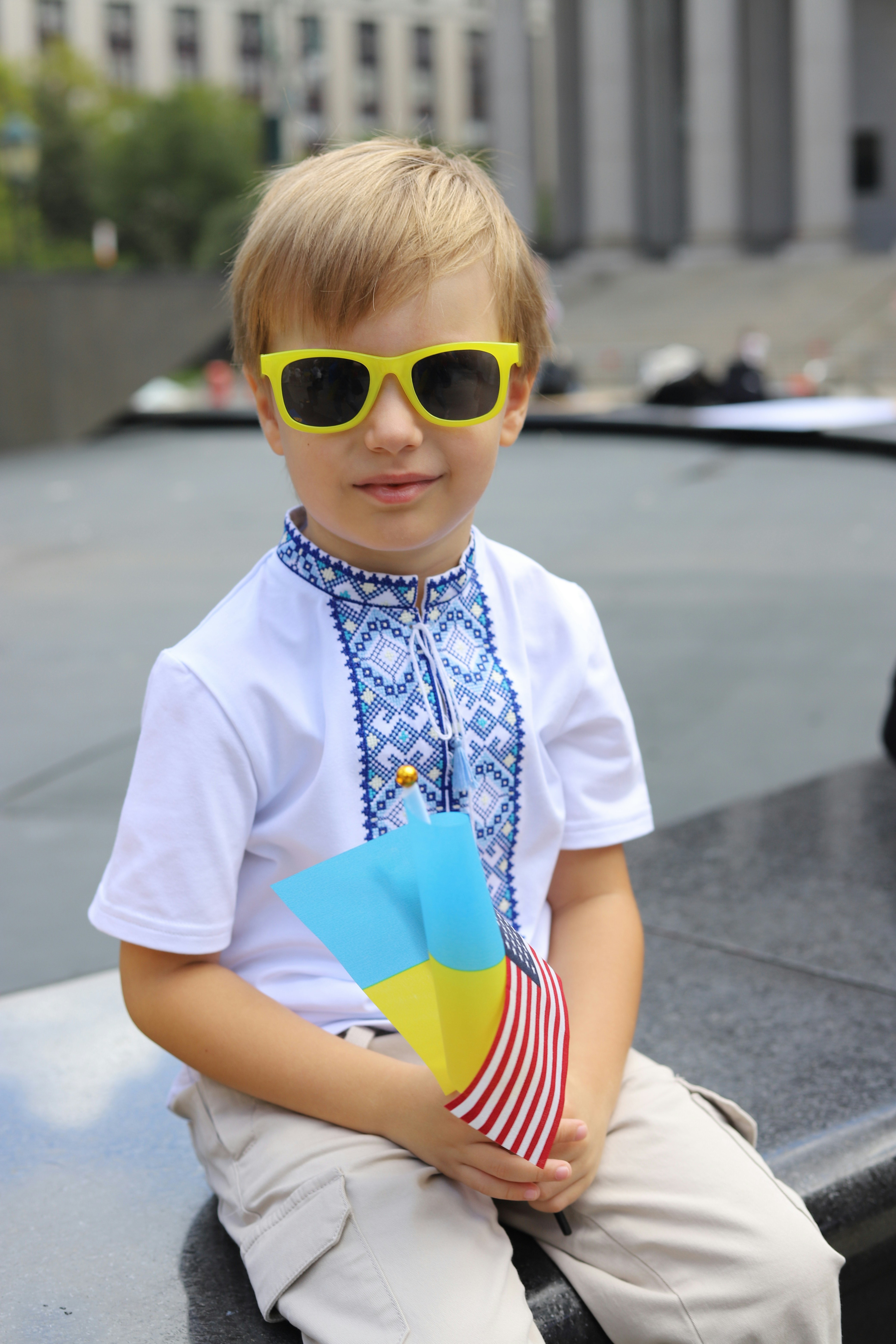 A child in yellow sunglasses and an embroidered shirt holding a small Ukrainian flag