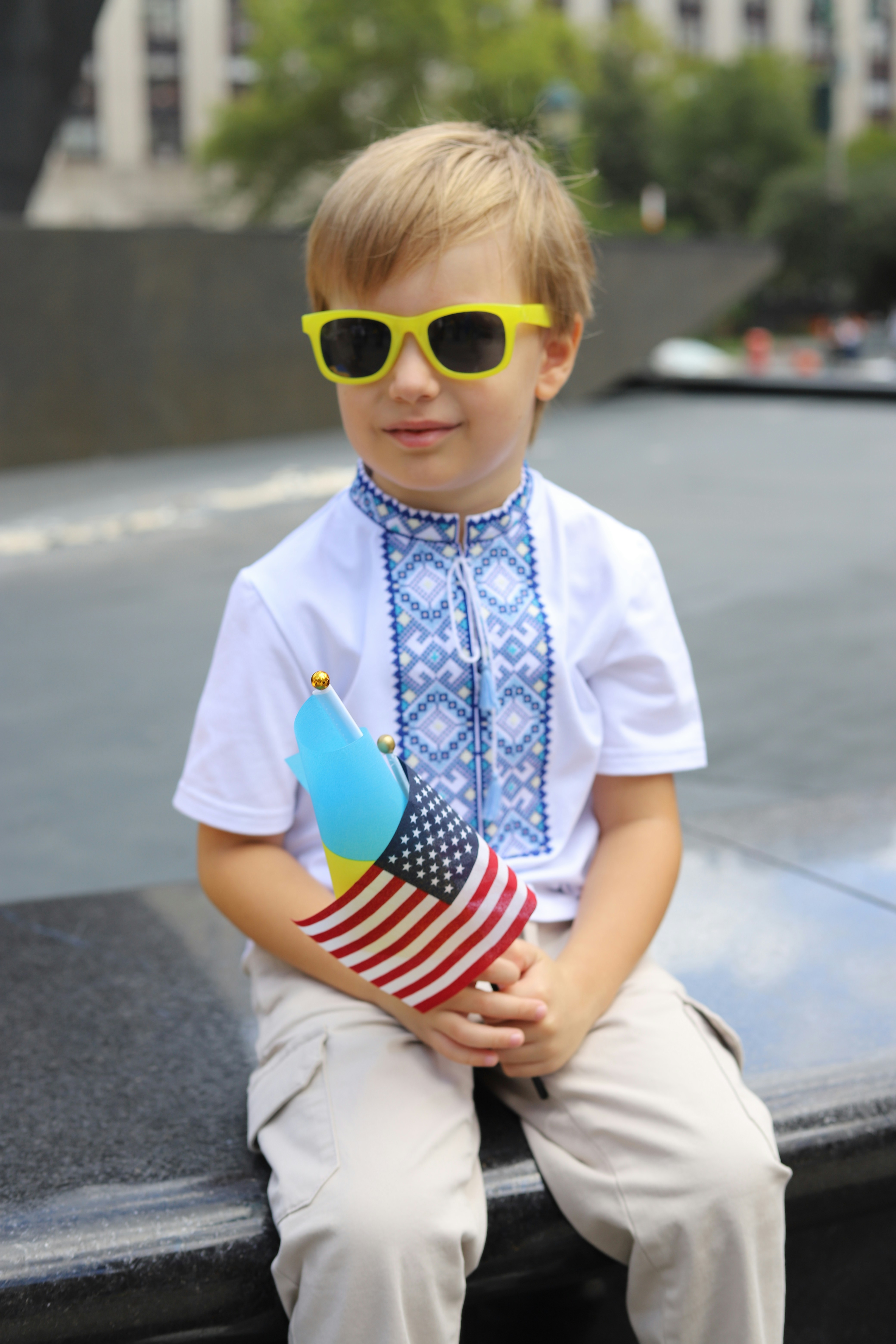 A child in a white embroidered shirt and yellow sunglasses holding a small American flag