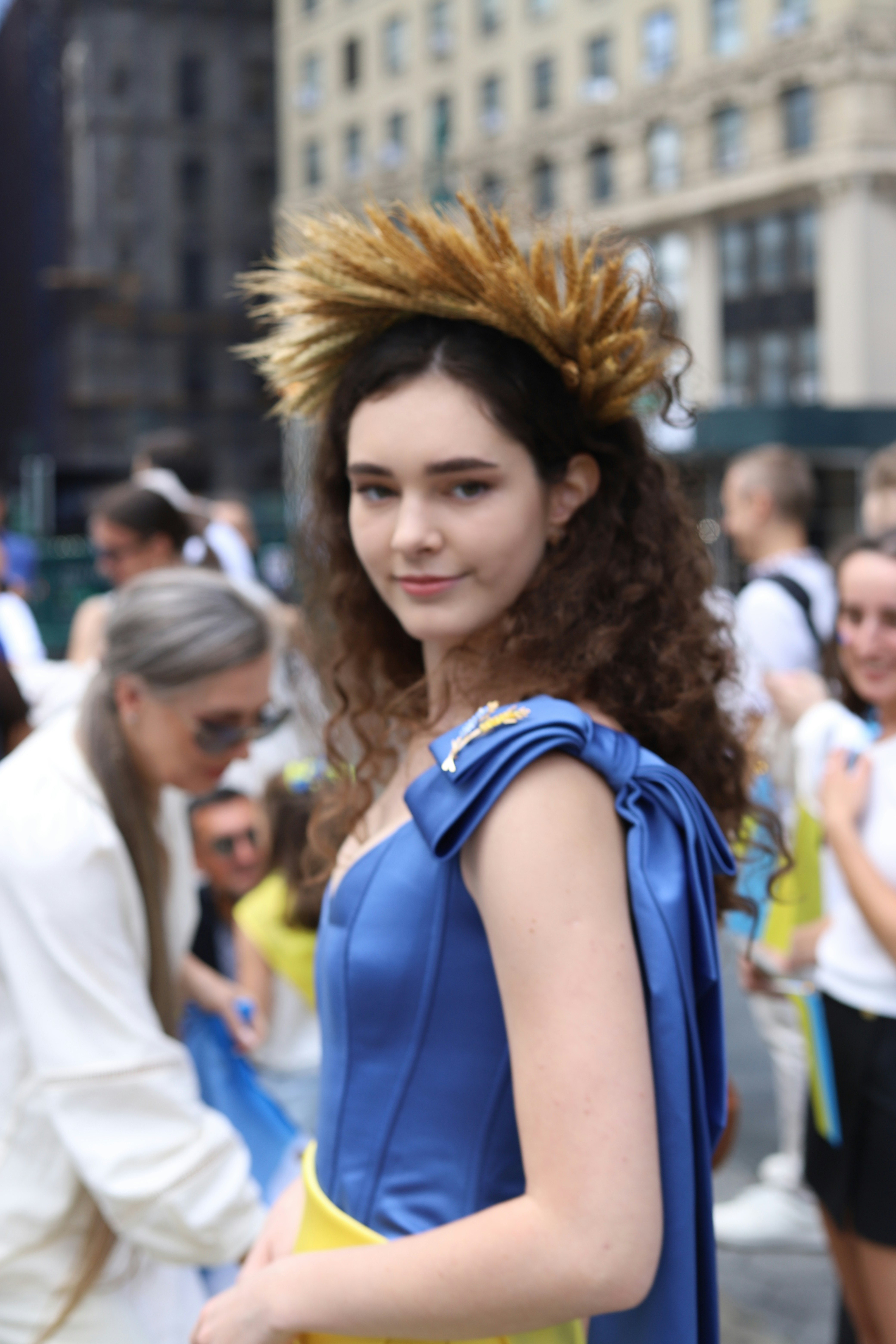 A young woman wearing a wheat crown and blue dress in a city crowd