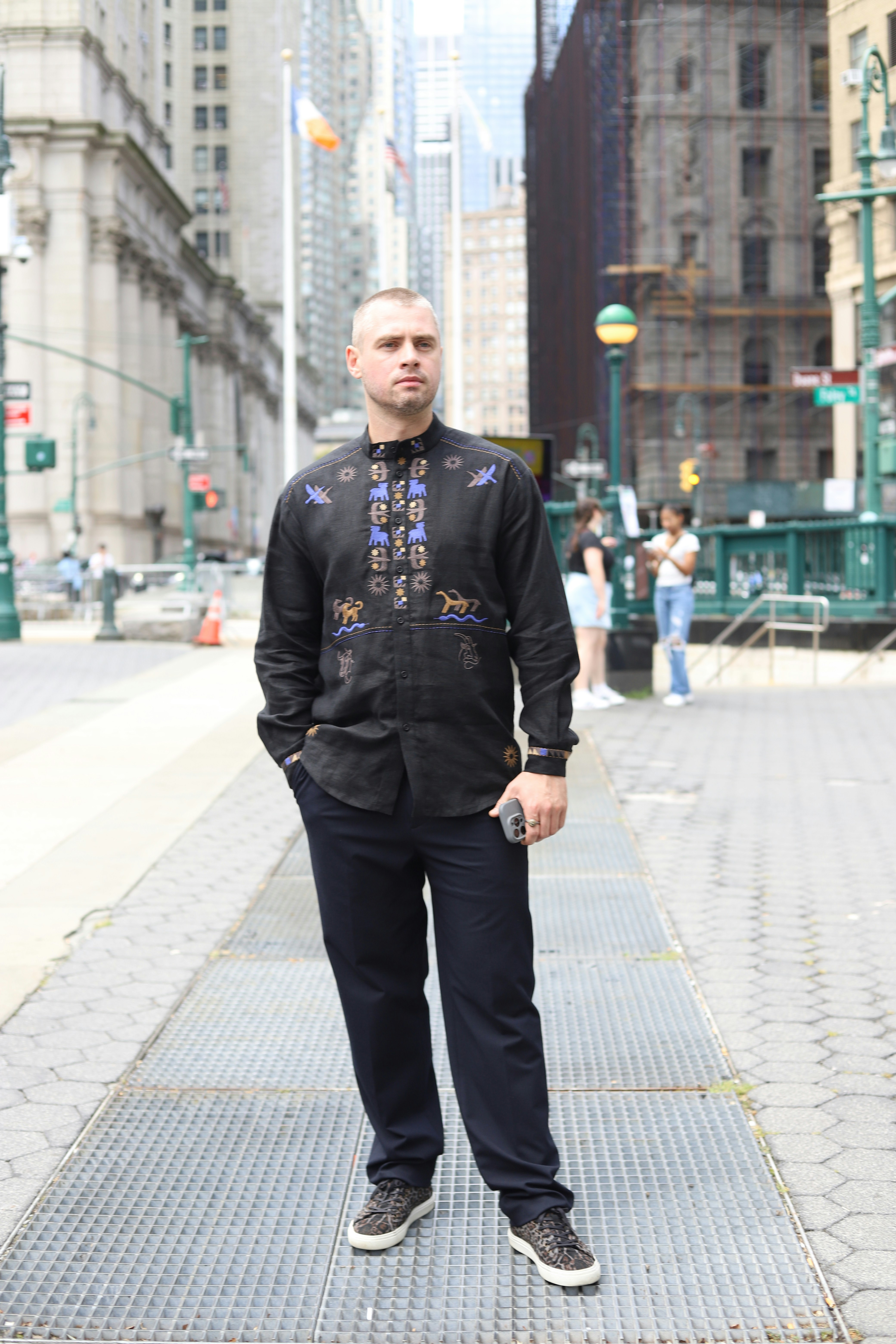 A man in a black embroidered shirt and dark trousers on a city sidewalk