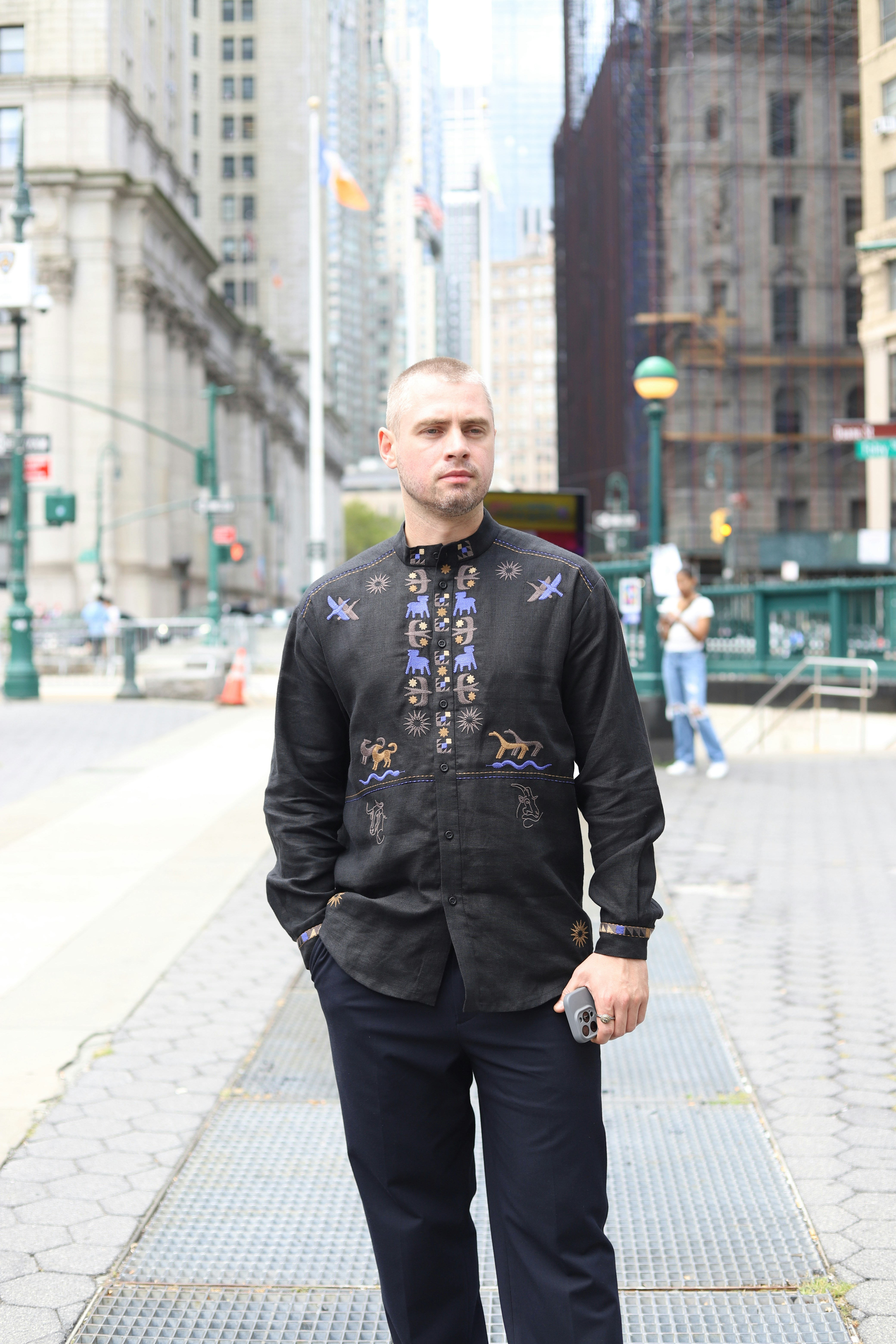 A man in a black embroidered shirt standing on a city sidewalk