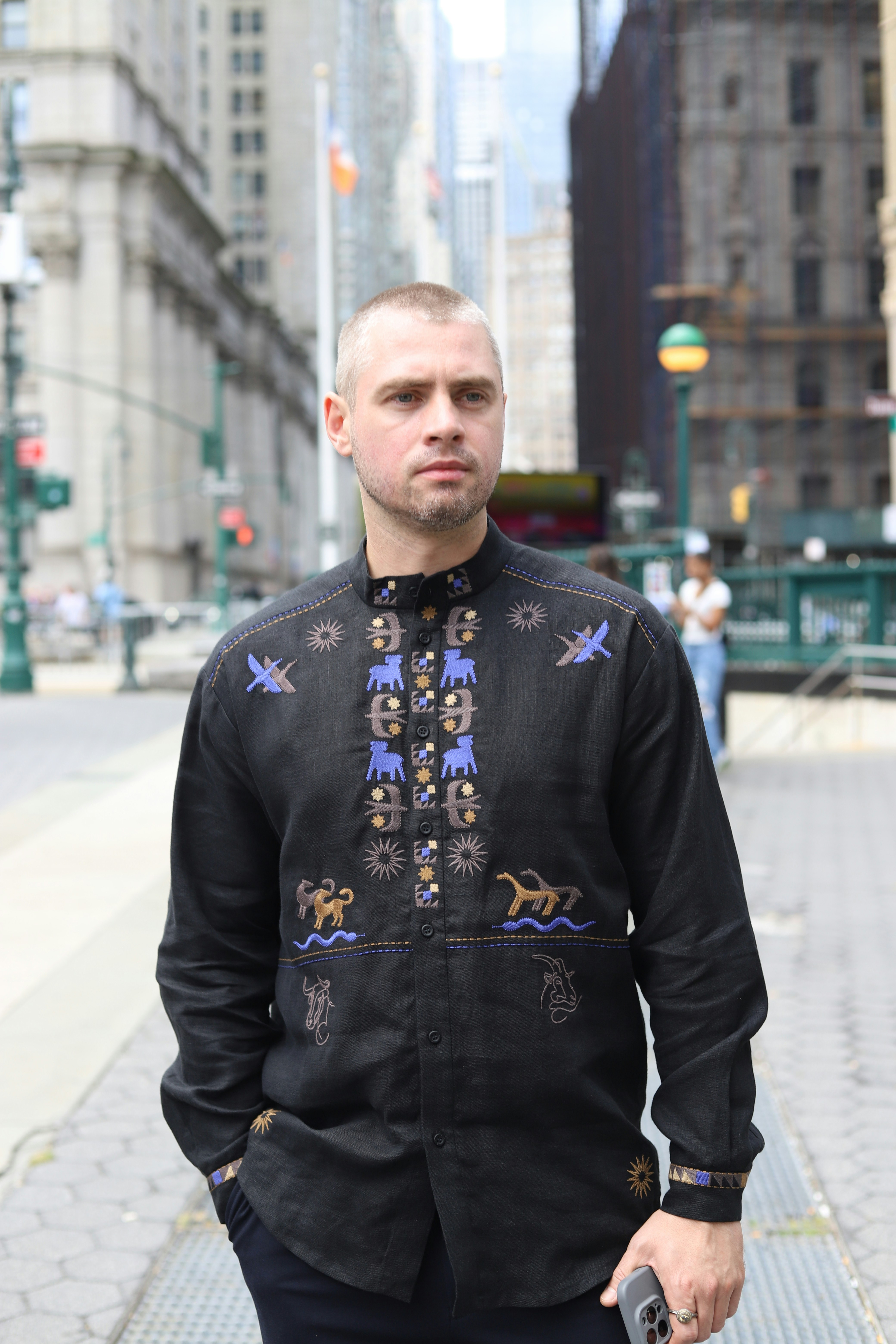 A man with short hair wearing a black embroidered shirt on a city street
