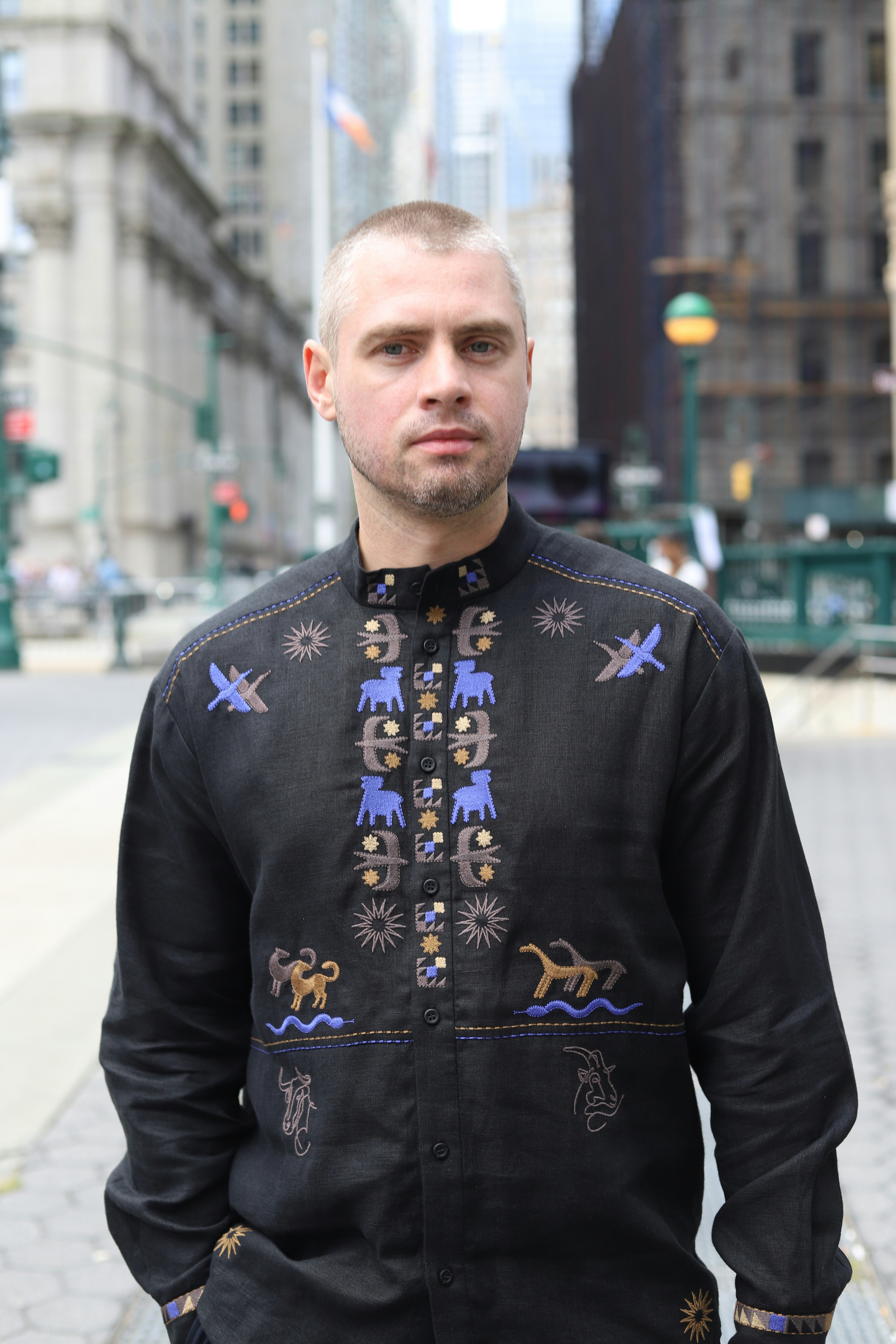 A man with short blonde hair wearing a black embroidered shirt on a city street