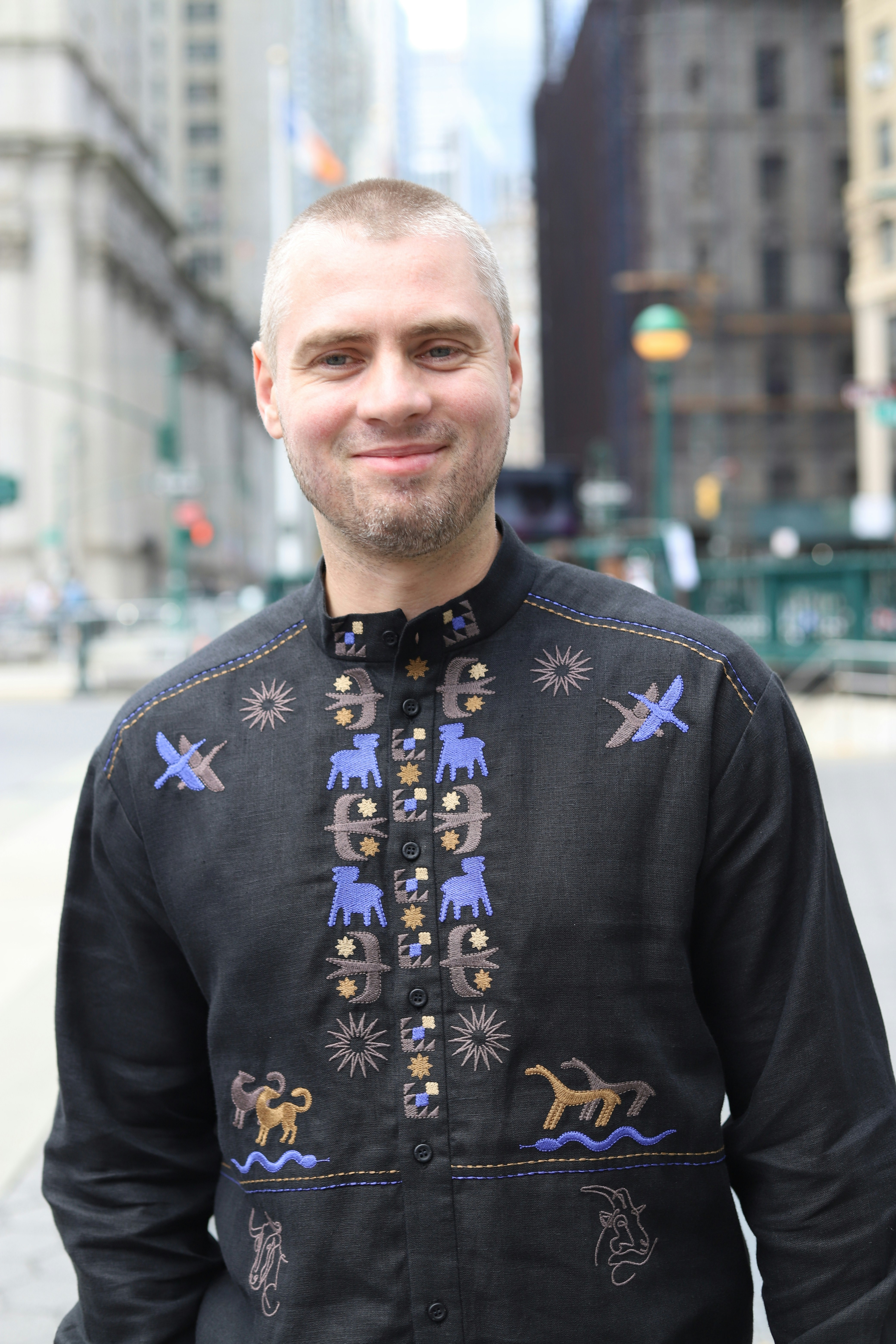 A man with short hair wearing a black embroidered shirt on a city street
