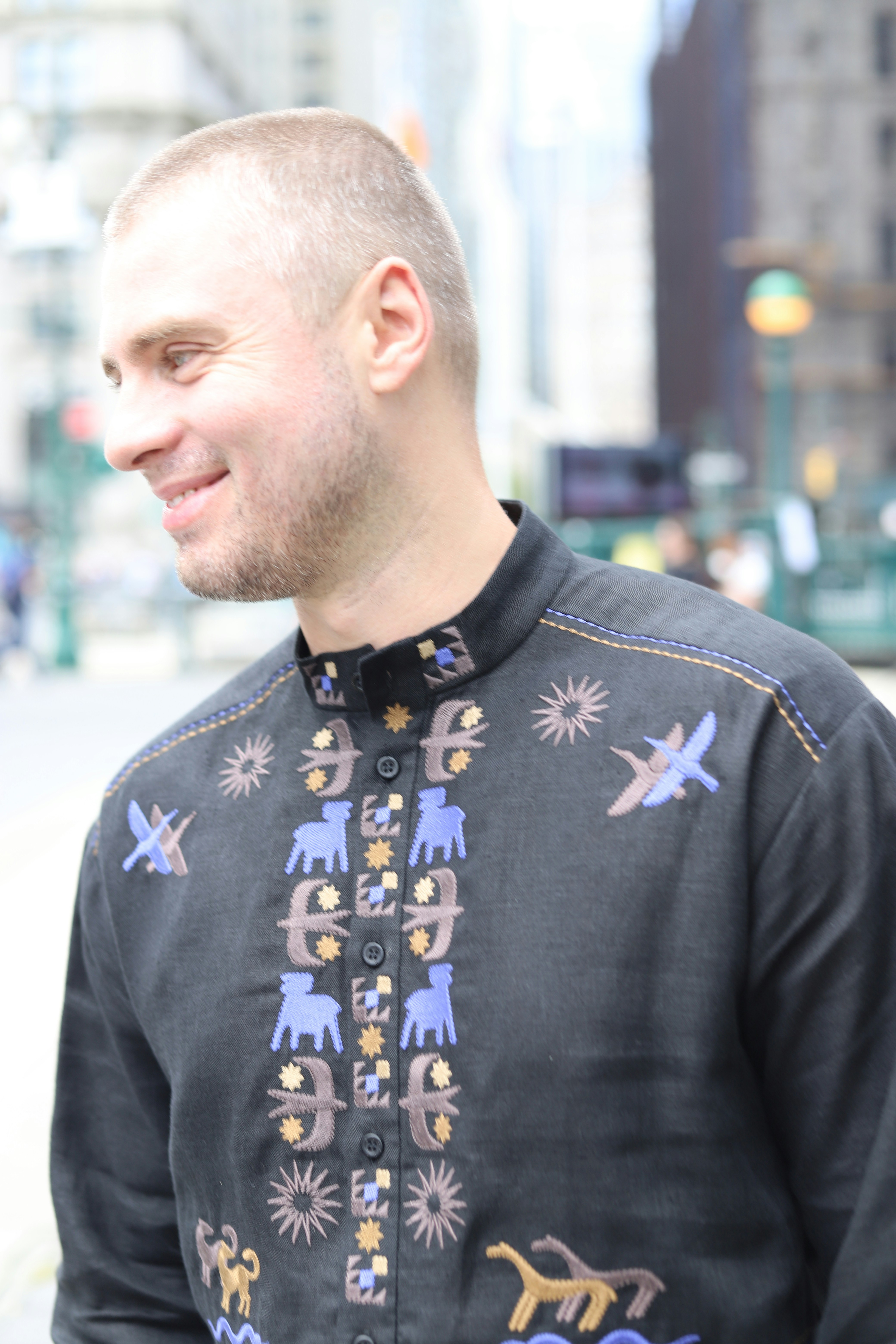 A man with a buzz cut wearing a black embroidered shirt on a city street