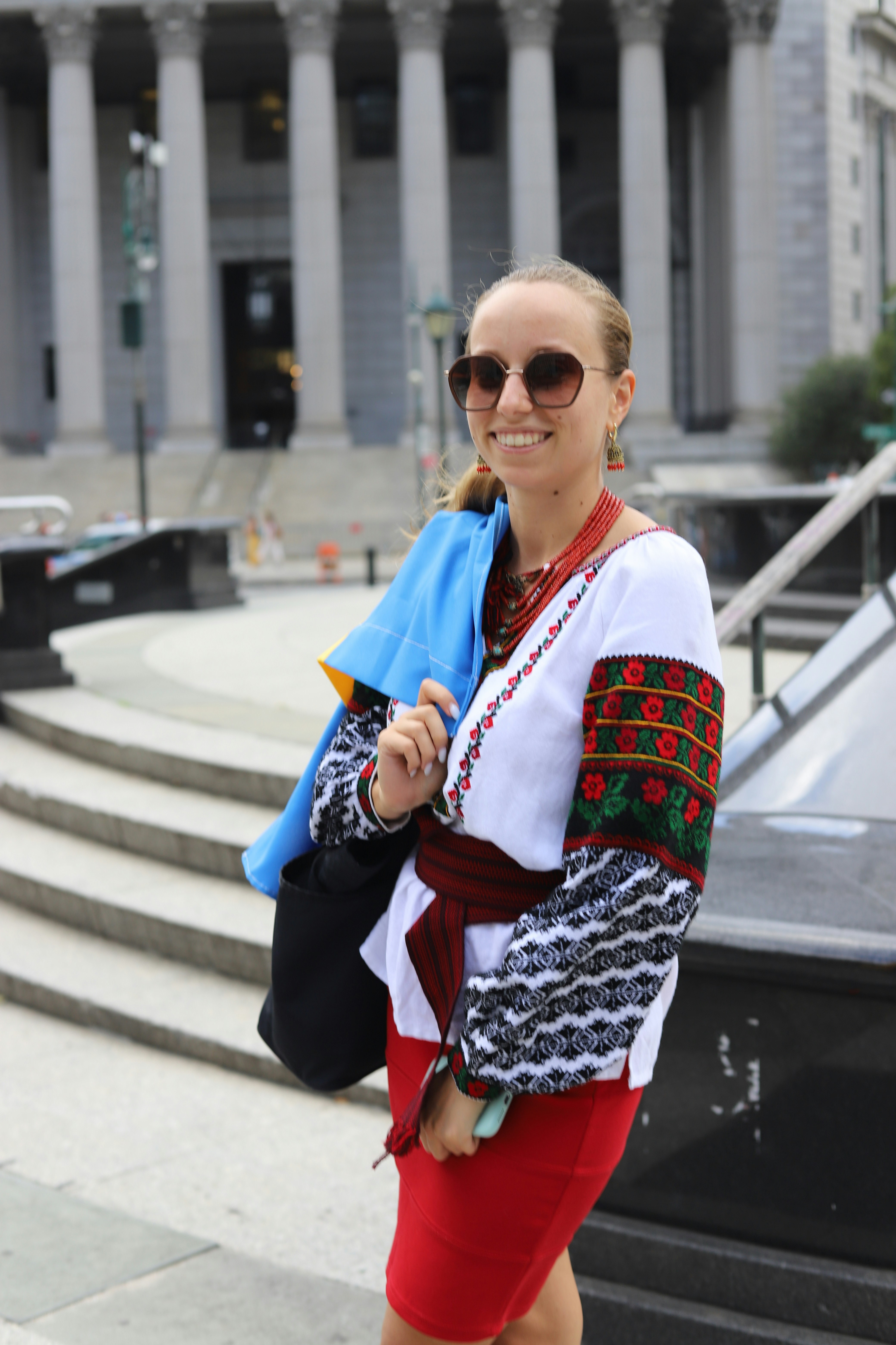 A woman wearing embroidered traditional clothing and sunglasses, holding a blue flag outdoors