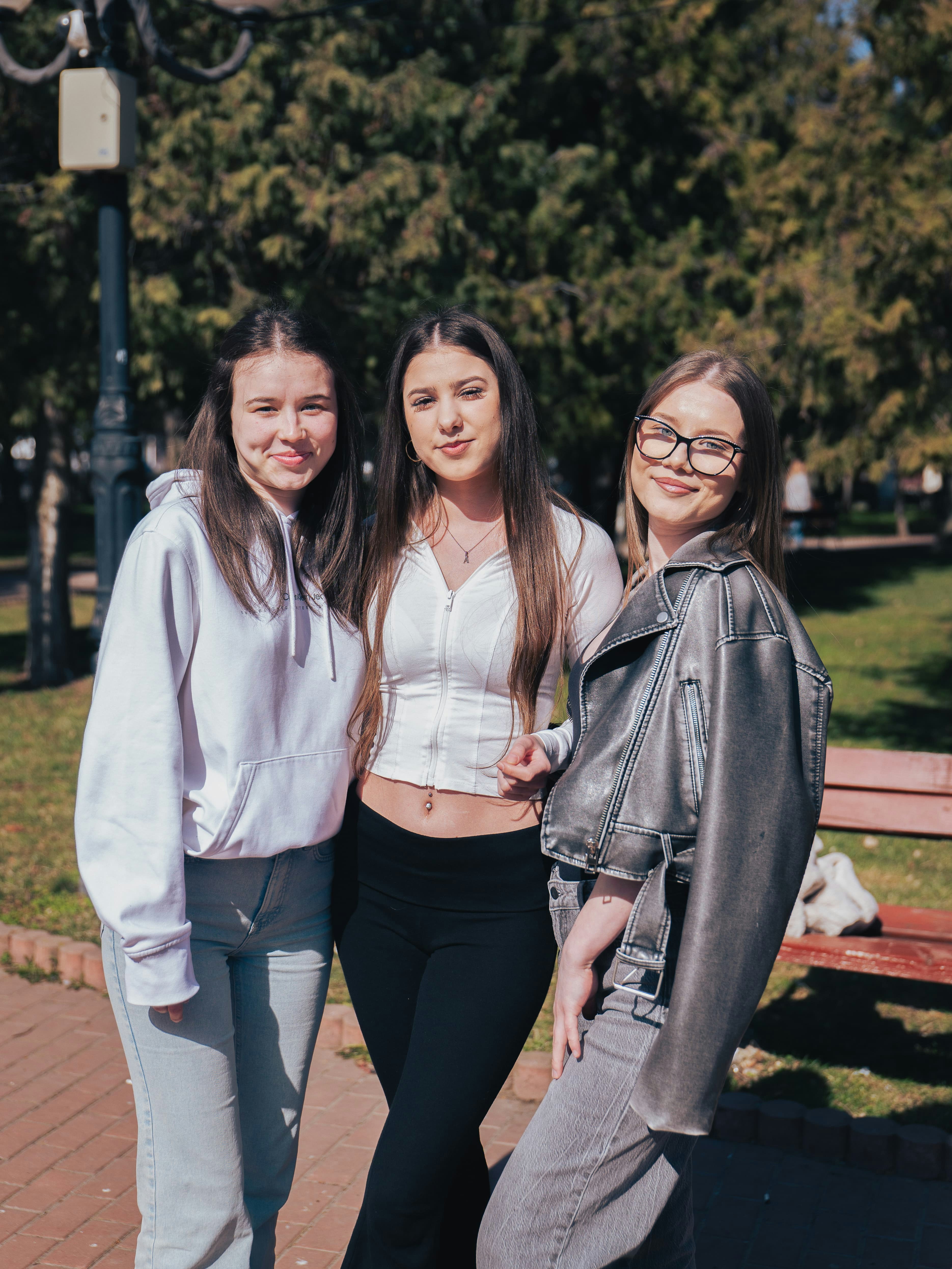 Three young women standing together in a sunlit park with trees in the background