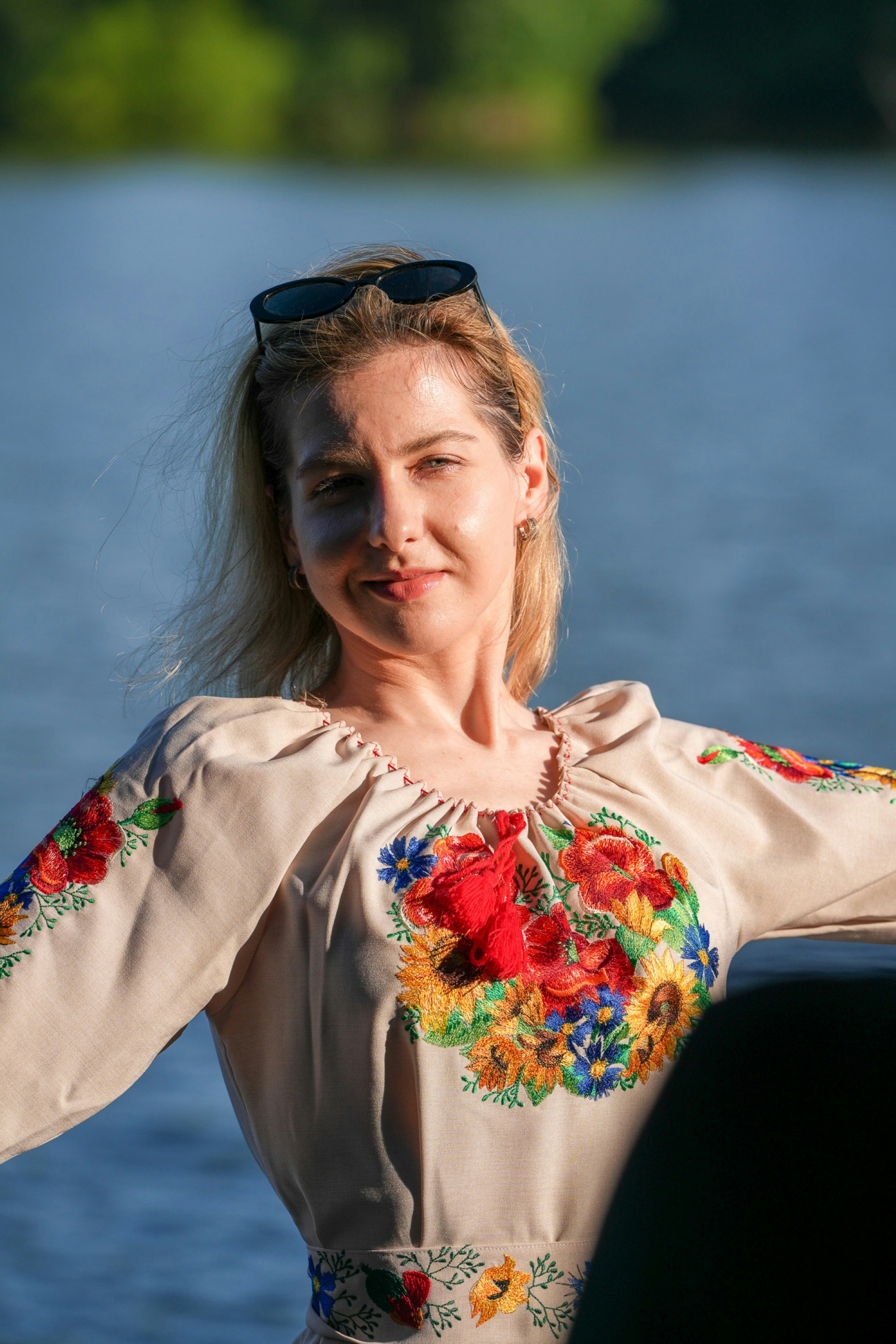 A woman wearing a beige embroidered blouse with floral patterns against a blue water background