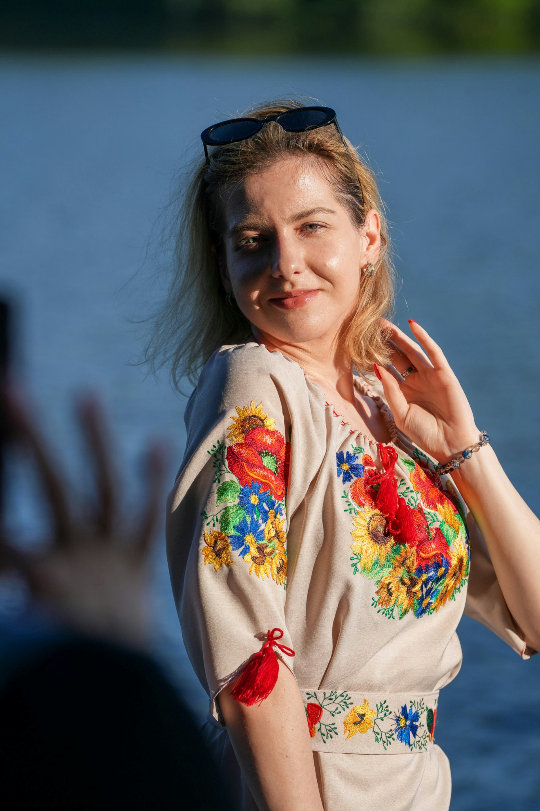 A woman wearing a beige embroidered blouse with floral patterns near a blue lake