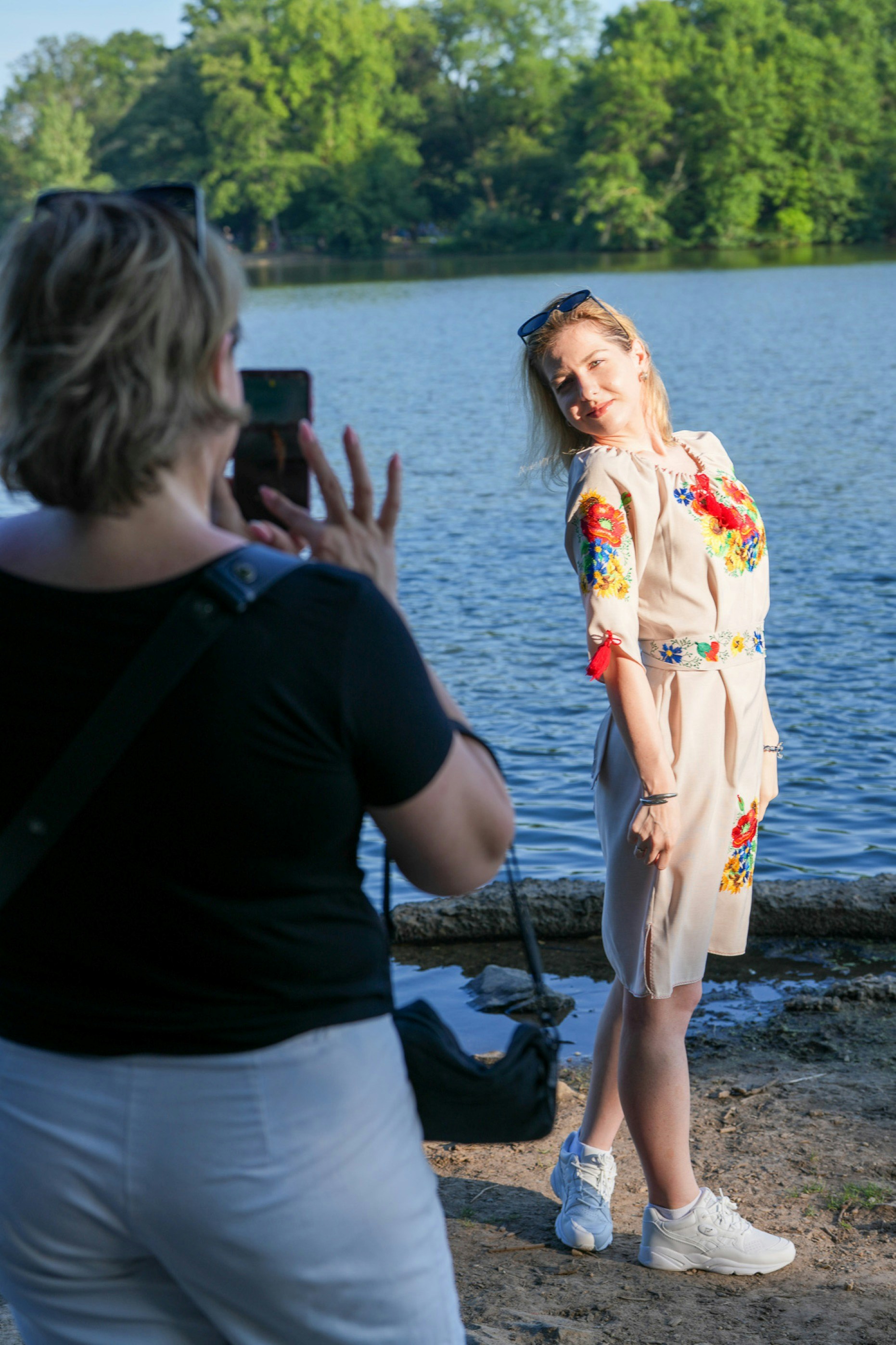 A woman photographing another woman in an embroidered dress by a lake