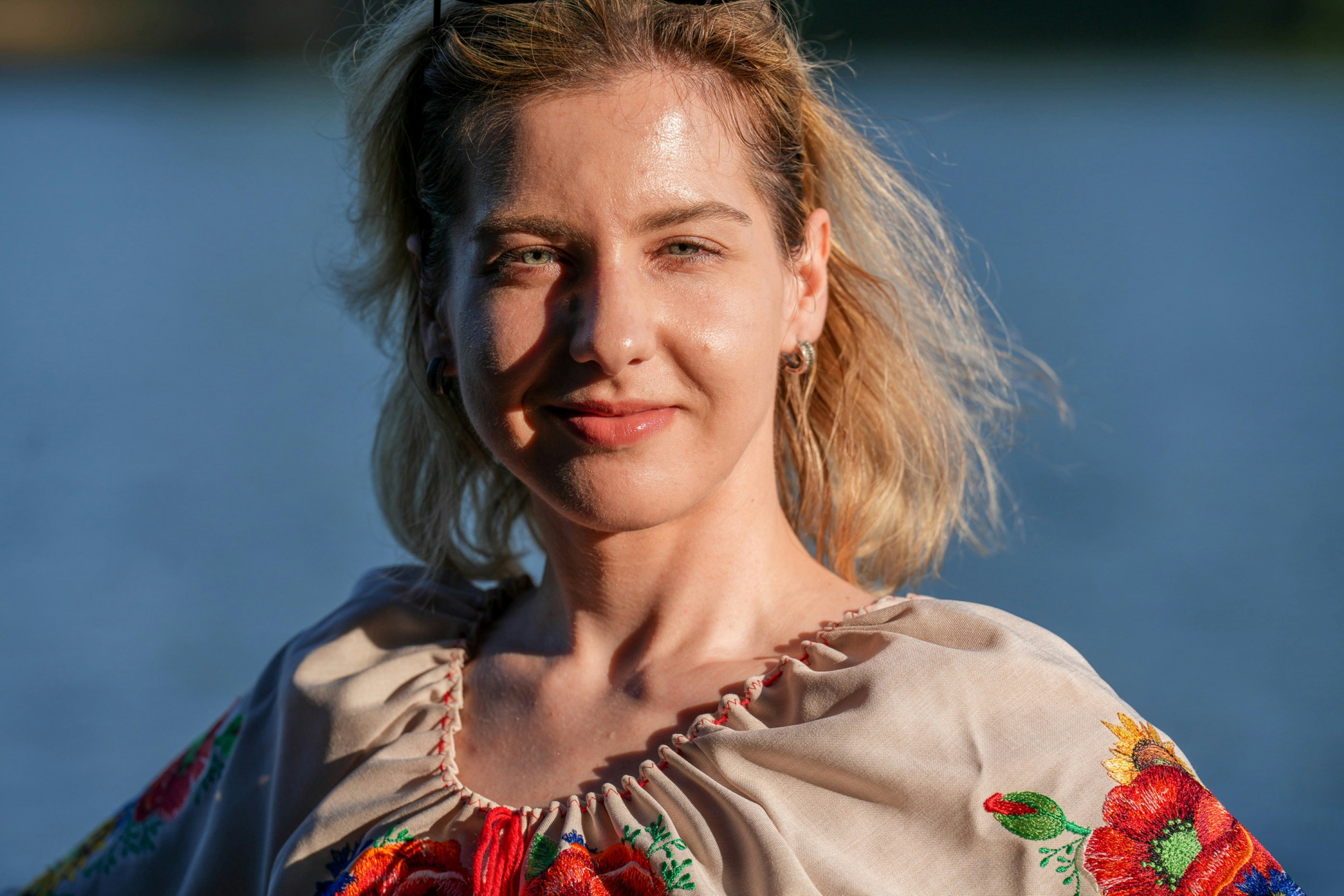 A woman in a beige embroidered blouse against a blurred blue water background
