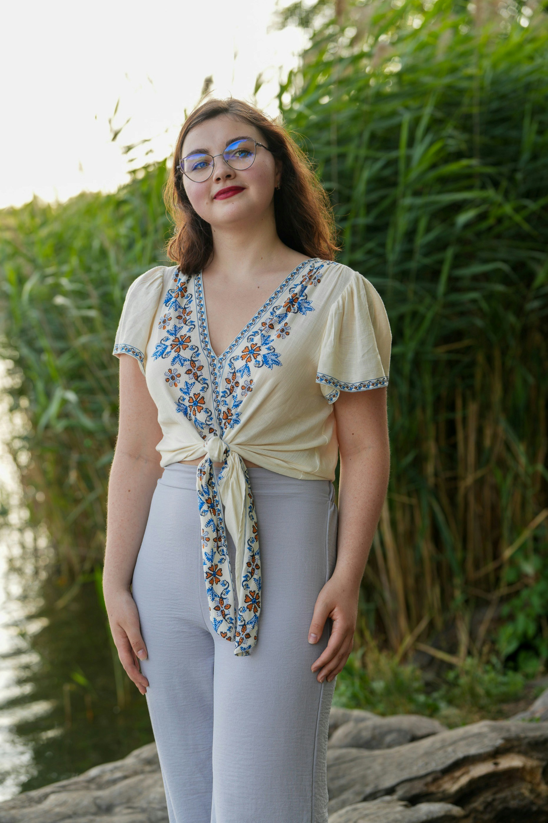 A woman in glasses and a floral tie-front top by a reedy lake