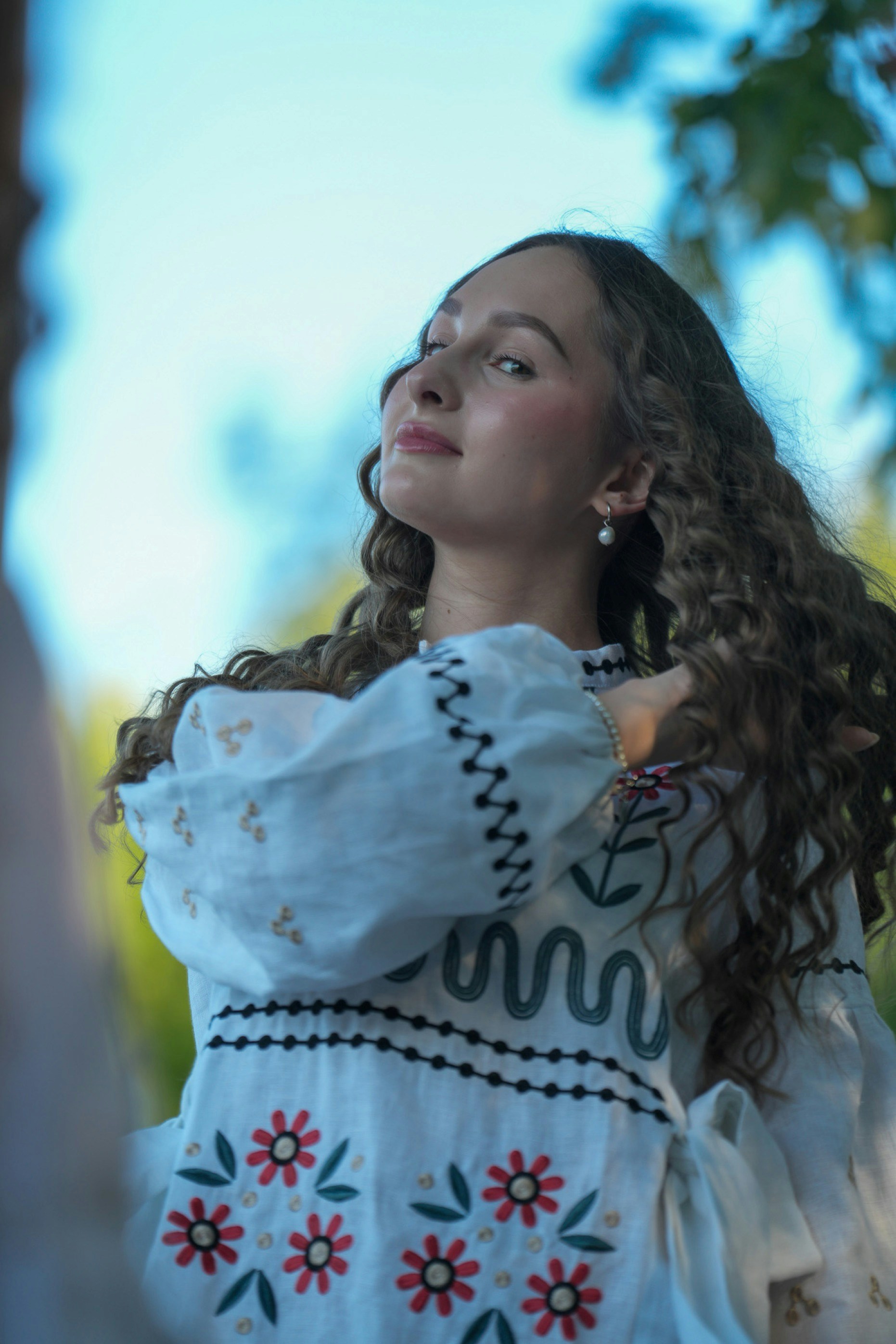 A woman with long curly hair wearing a white embroidered blouse outdoors