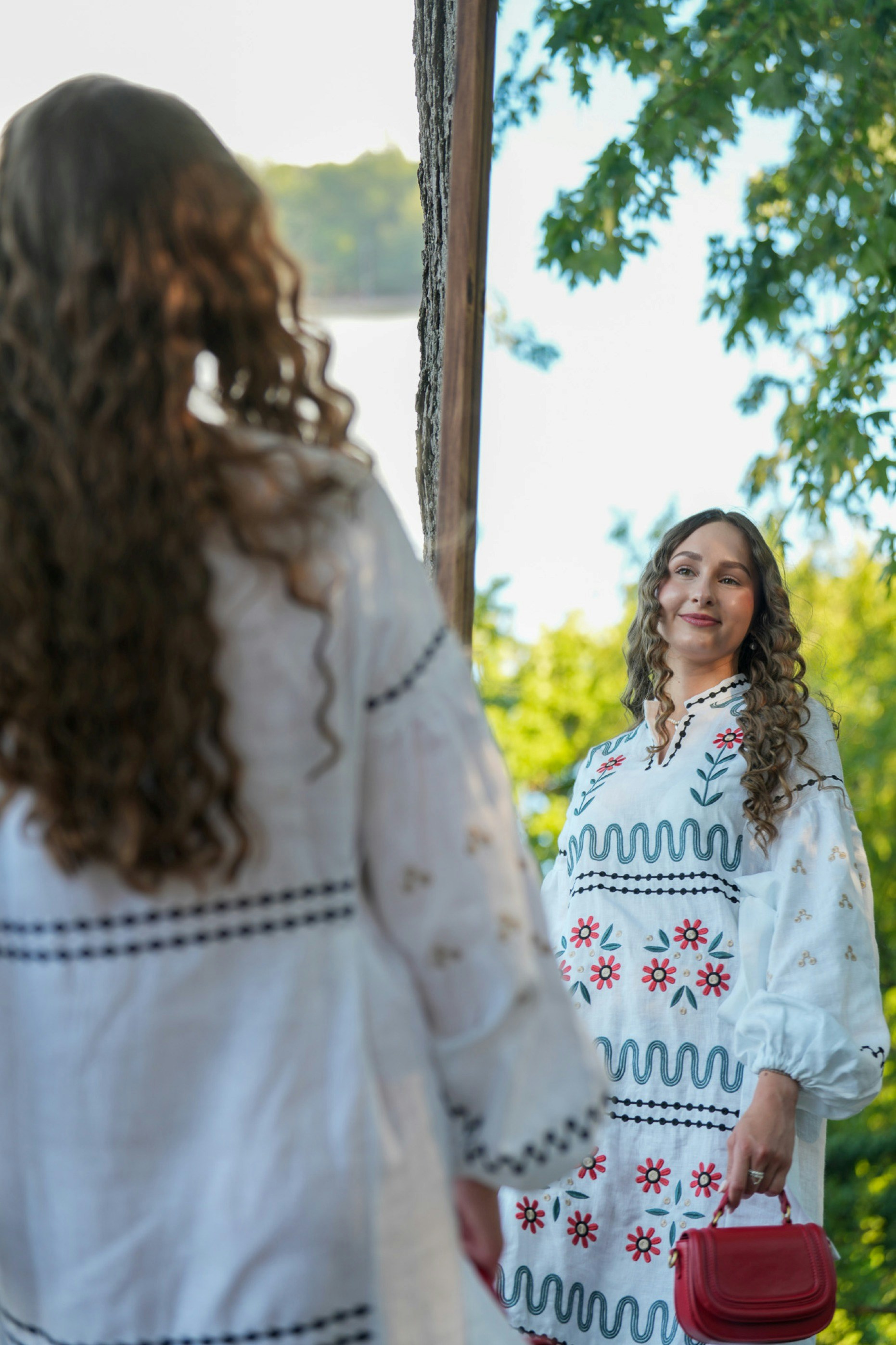 A woman with curly hair wearing a white embroidered dress looking into a mirror