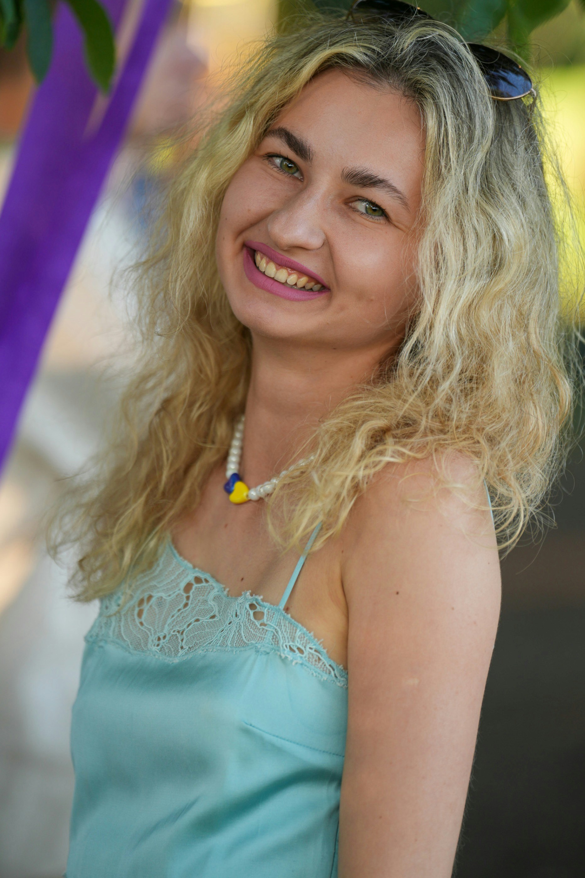 A smiling woman with blonde curly hair wearing a light blue lace camisole