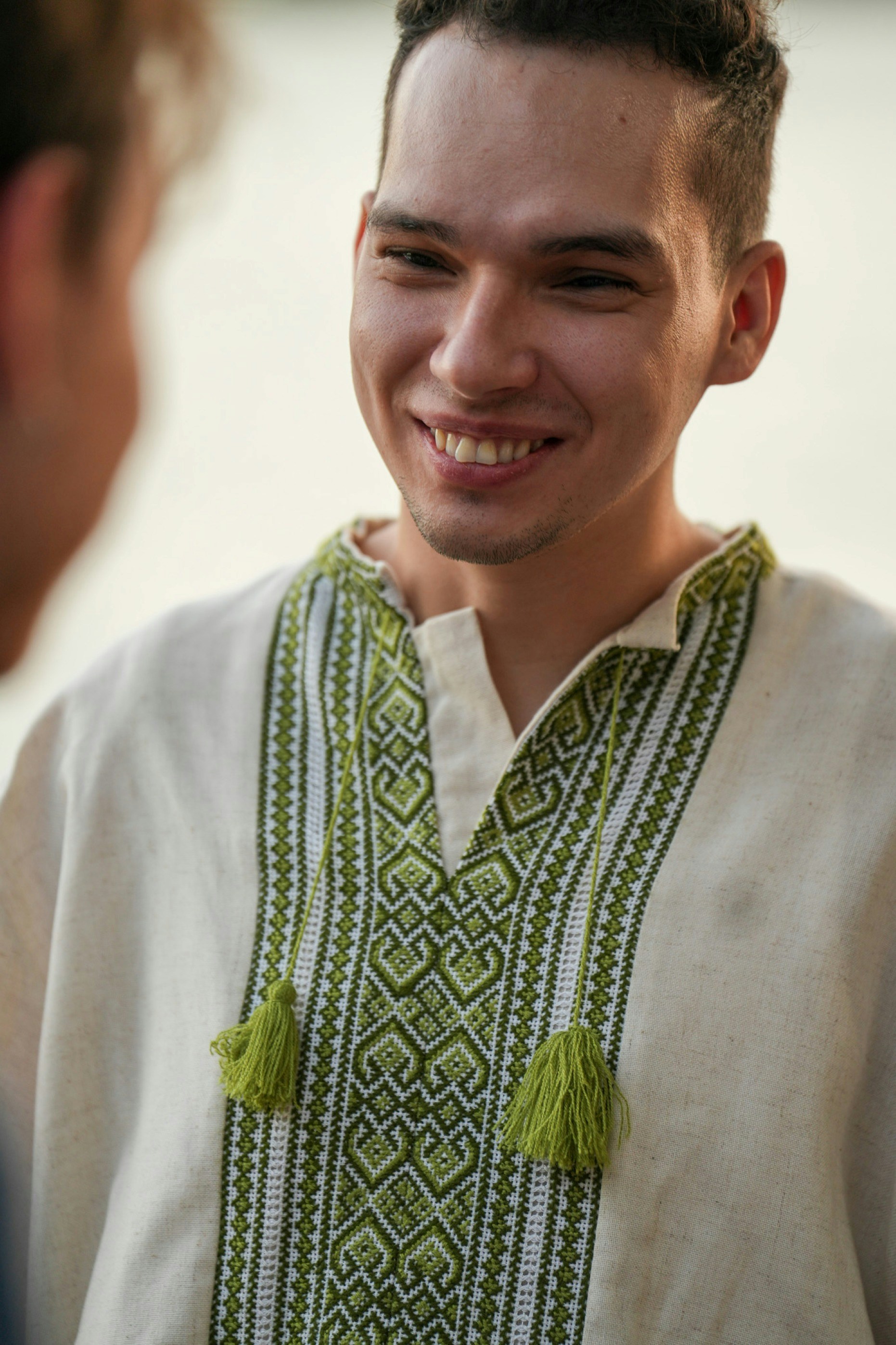 A smiling man wearing a cream-colored shirt with green geometric embroidery and tassels