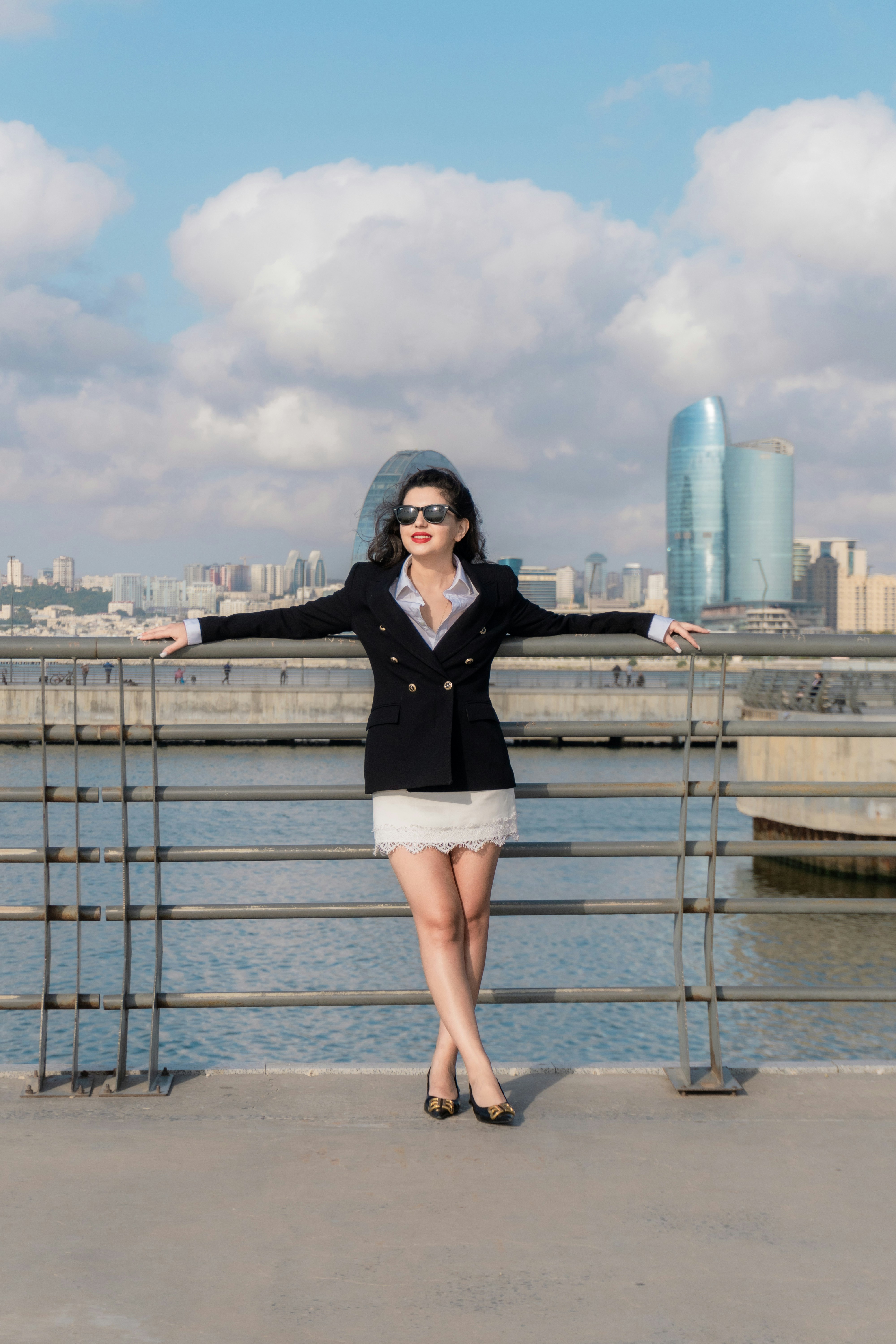 A woman in a black blazer and white skirt leaning against a metal railing