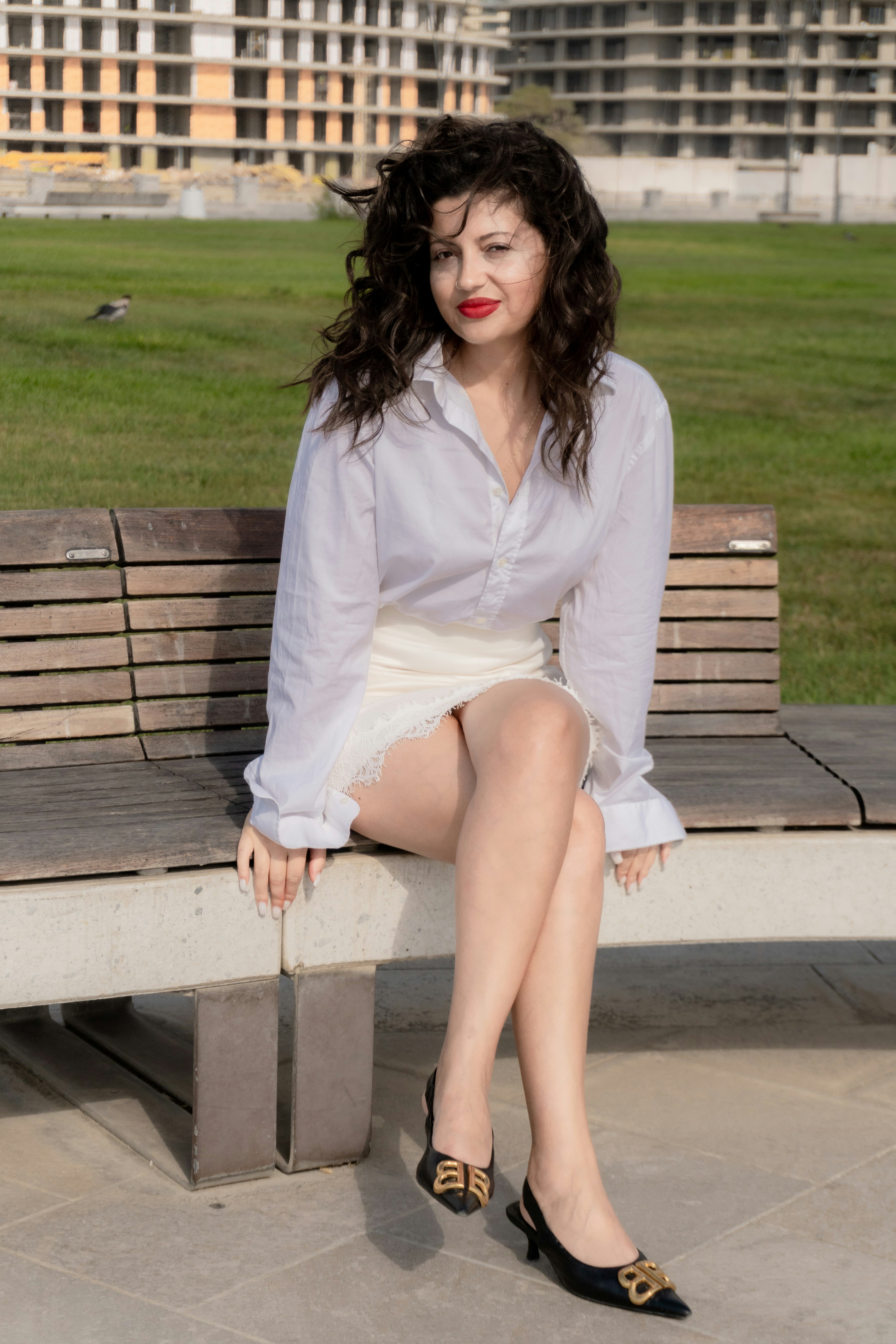 A woman with dark wavy hair in a white shirt and skirt on a bench