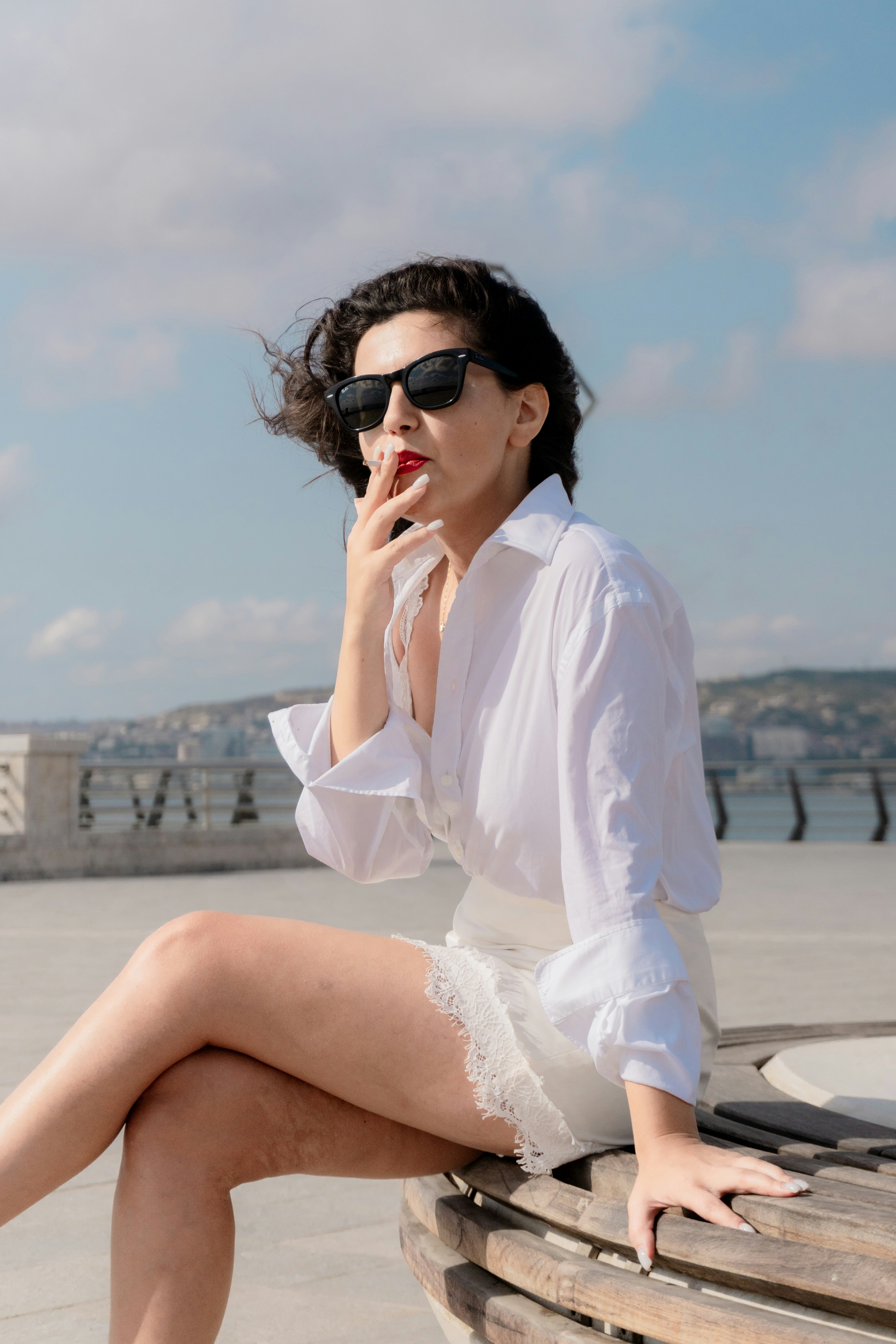 A woman in a white shirt and sunglasses smoking on a wooden bench