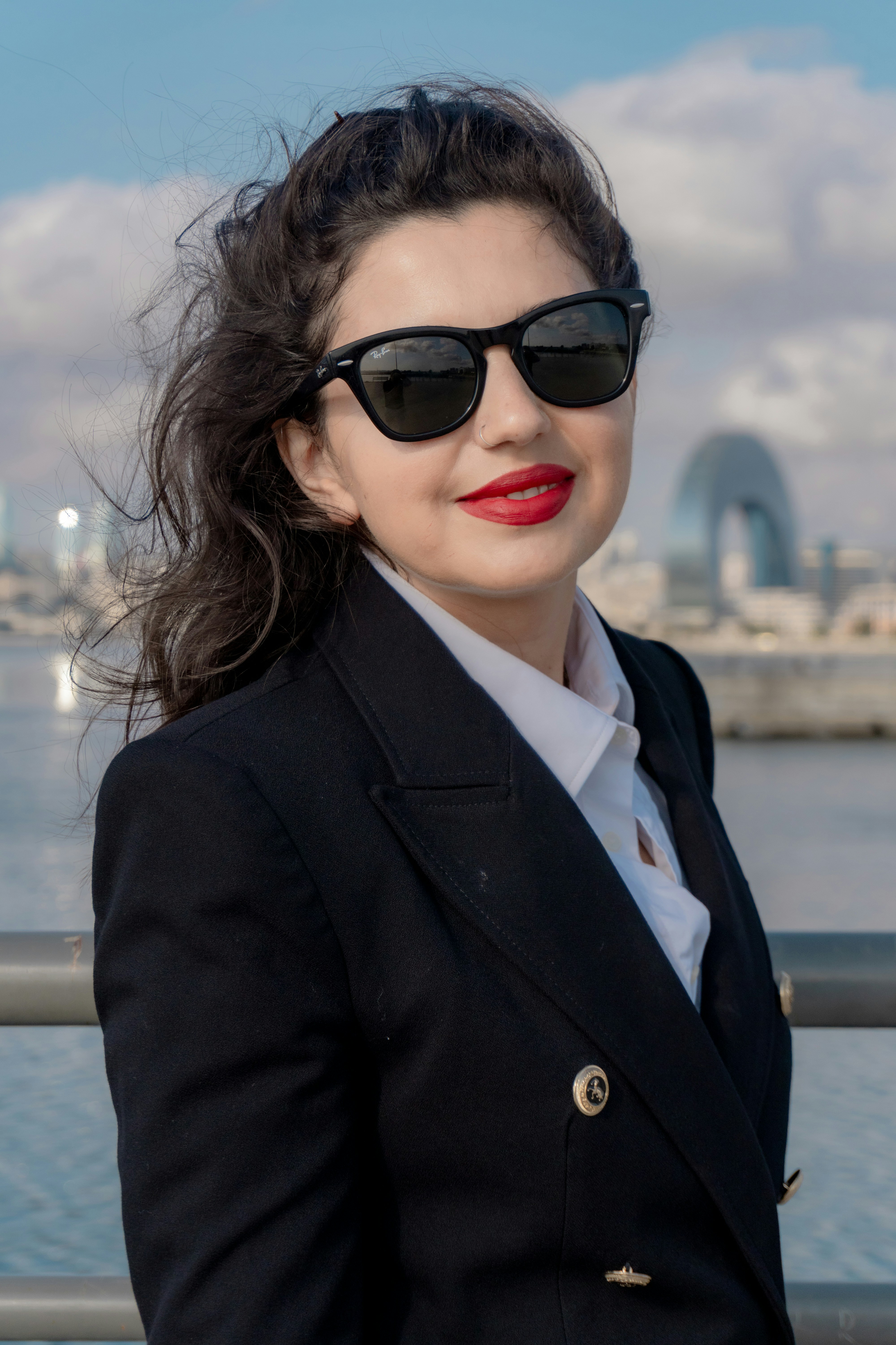 A woman in a black blazer and white shirt wearing sunglasses by the water