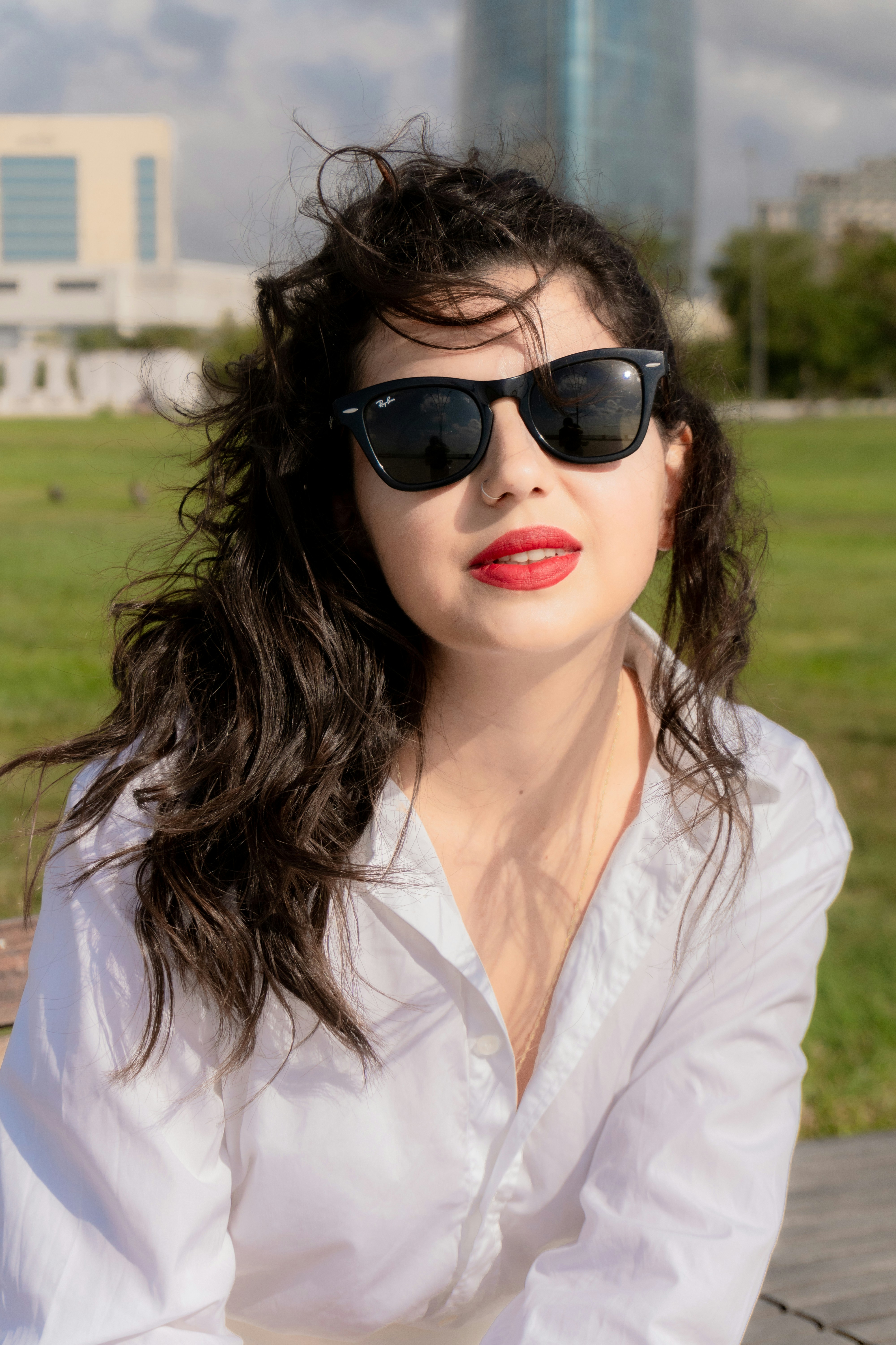 A woman with dark wavy hair wearing black sunglasses and a white shirt outdoors