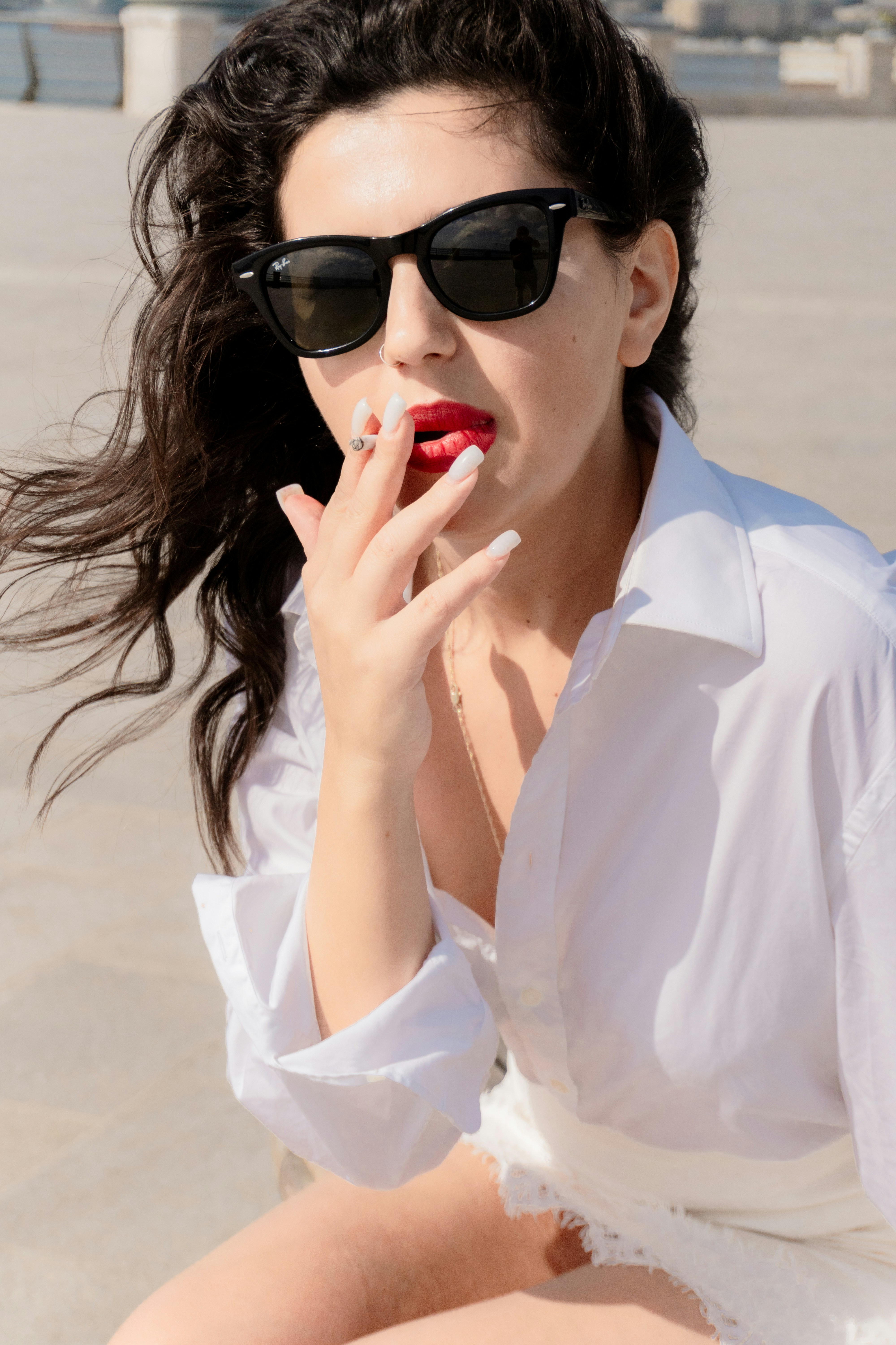 A woman with dark hair wearing sunglasses and a white shirt smoking a cigarette
