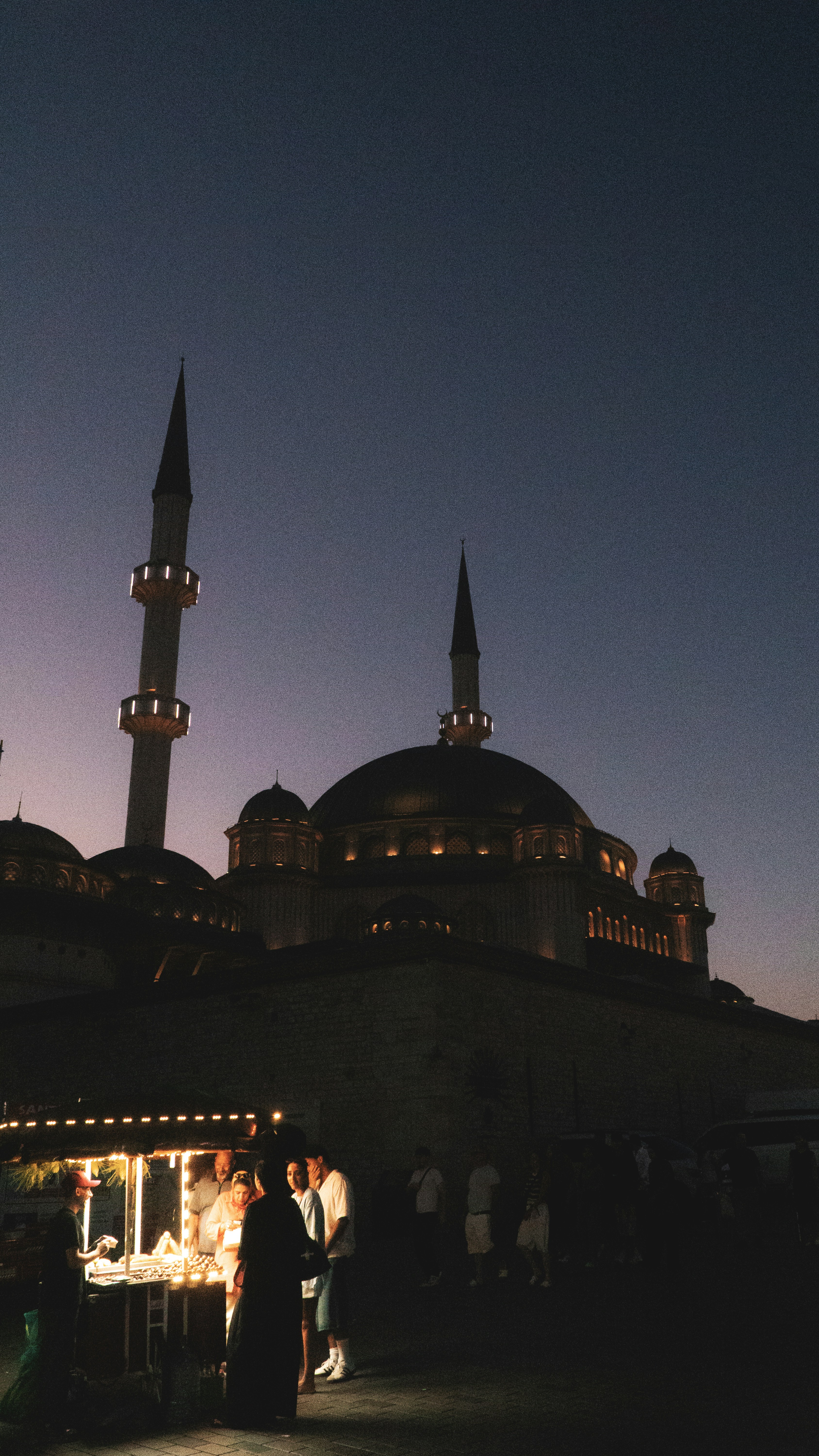 A food stall illuminated at night in front of a large mosque with minarets