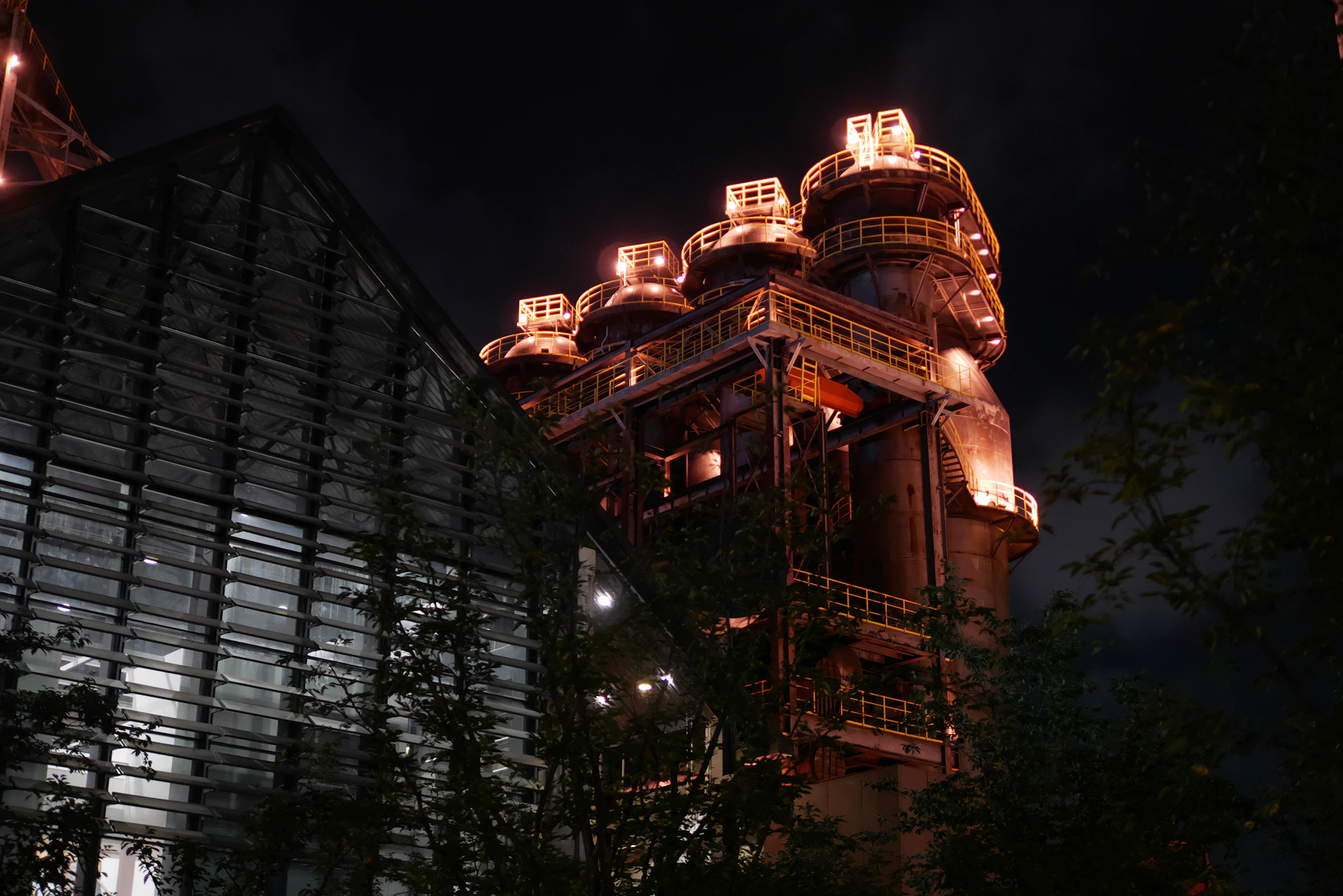 An illuminated industrial structure with tall towers at night beside a glass building