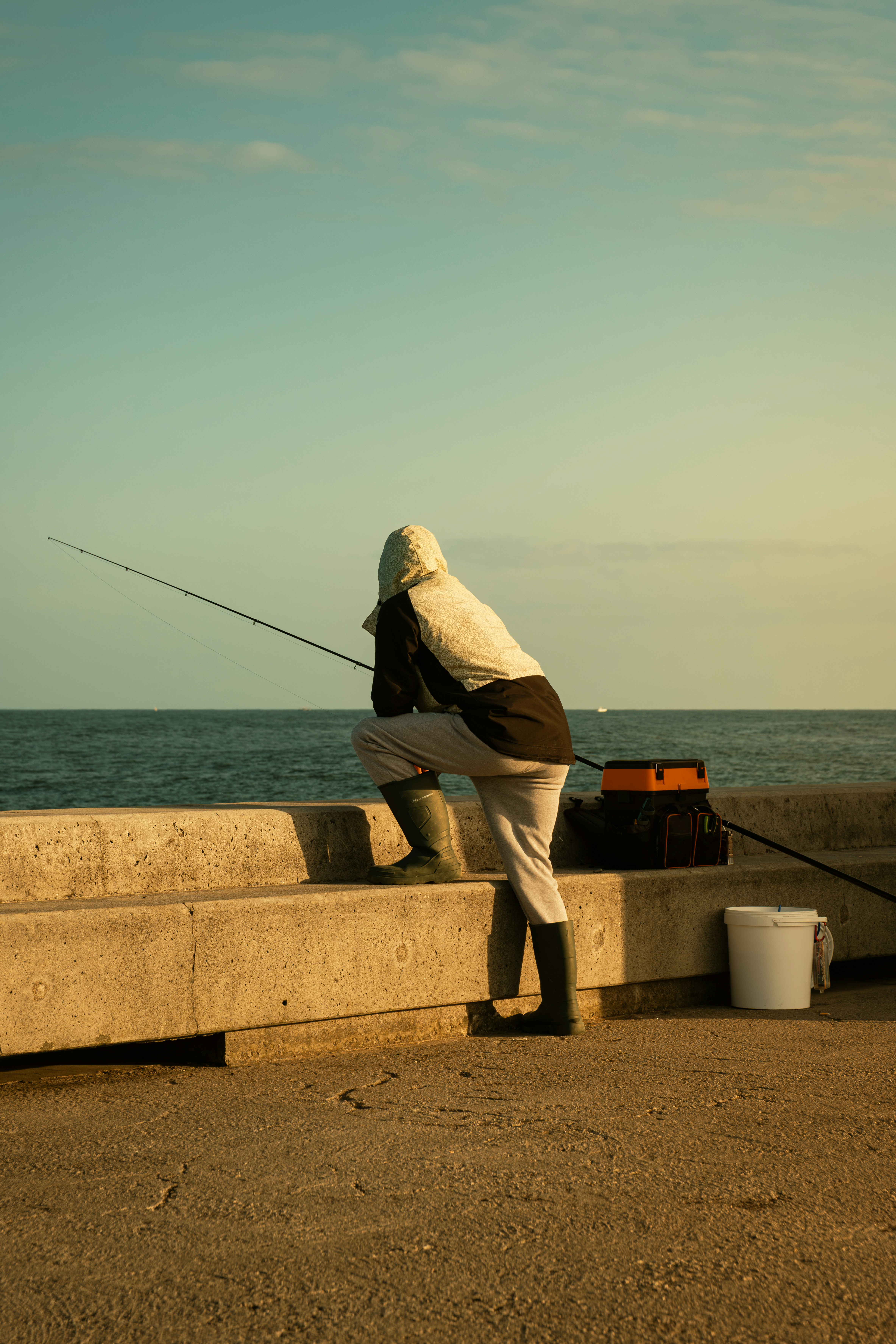 Hooded angler crouched on a concrete pier beside a rod and an orange tackle box, a flat sea behind