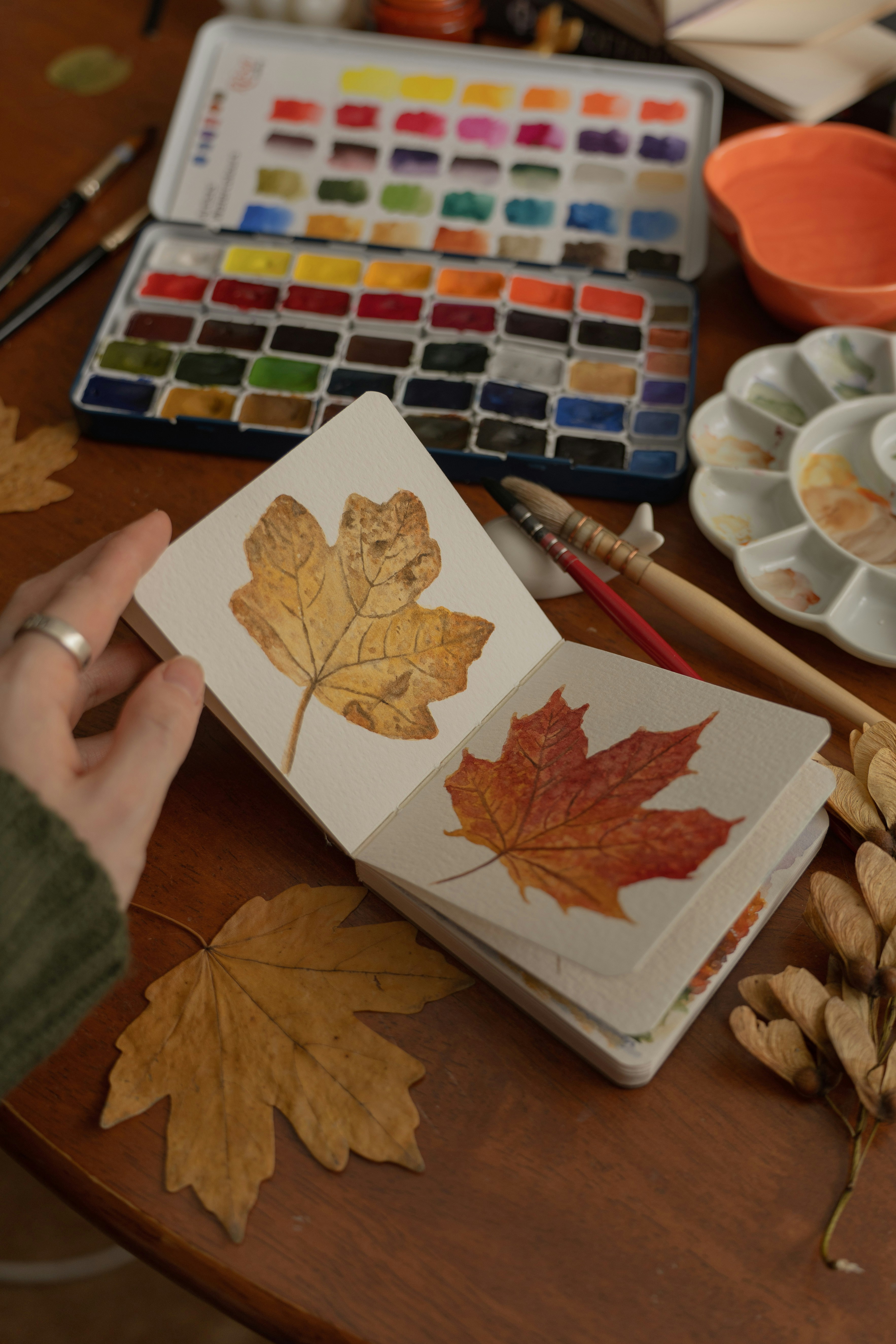 A hand holding a sketchbook with watercolor paintings of autumn leaves on a desk