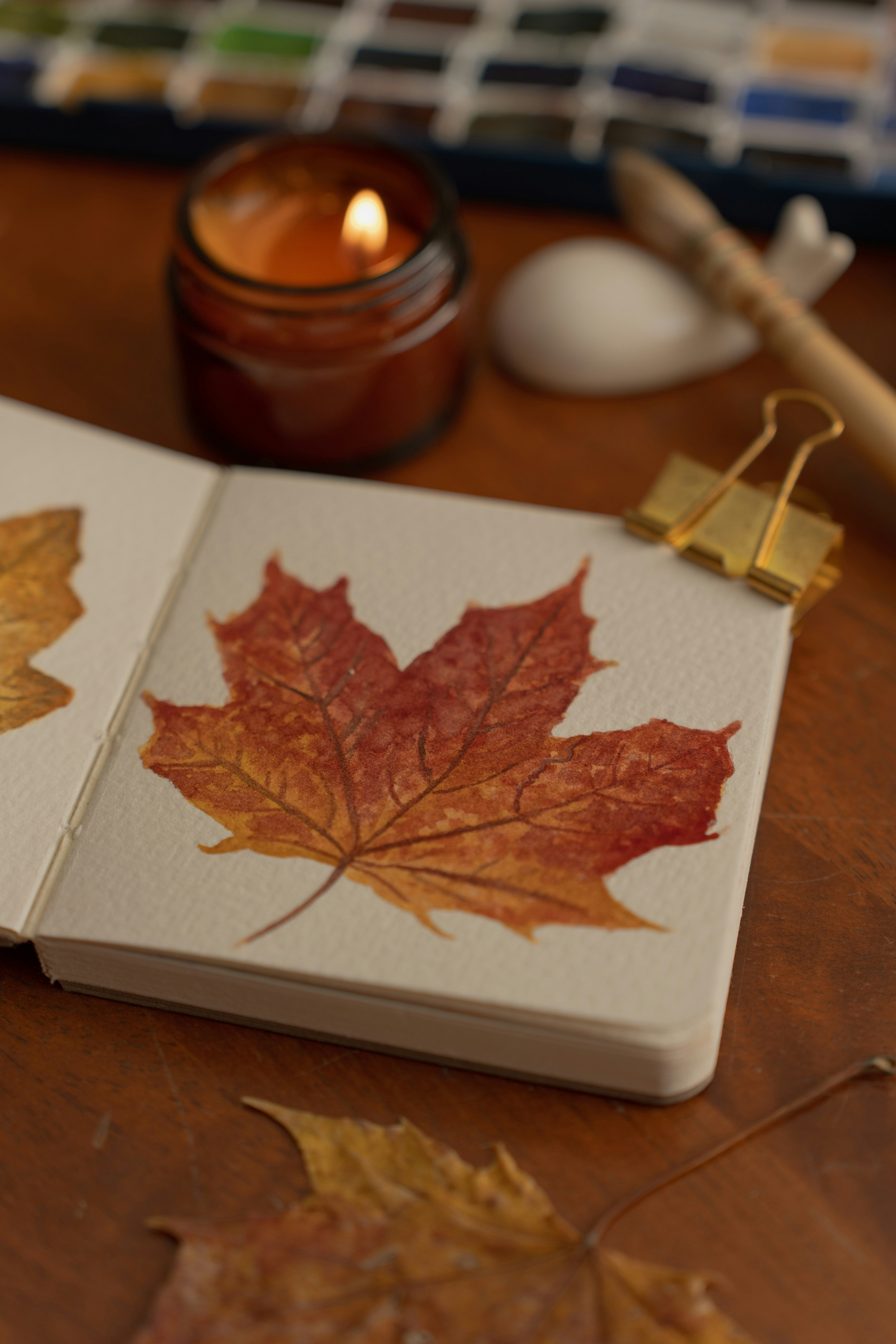 A watercolor painting of a maple leaf in an open sketchbook with a candle