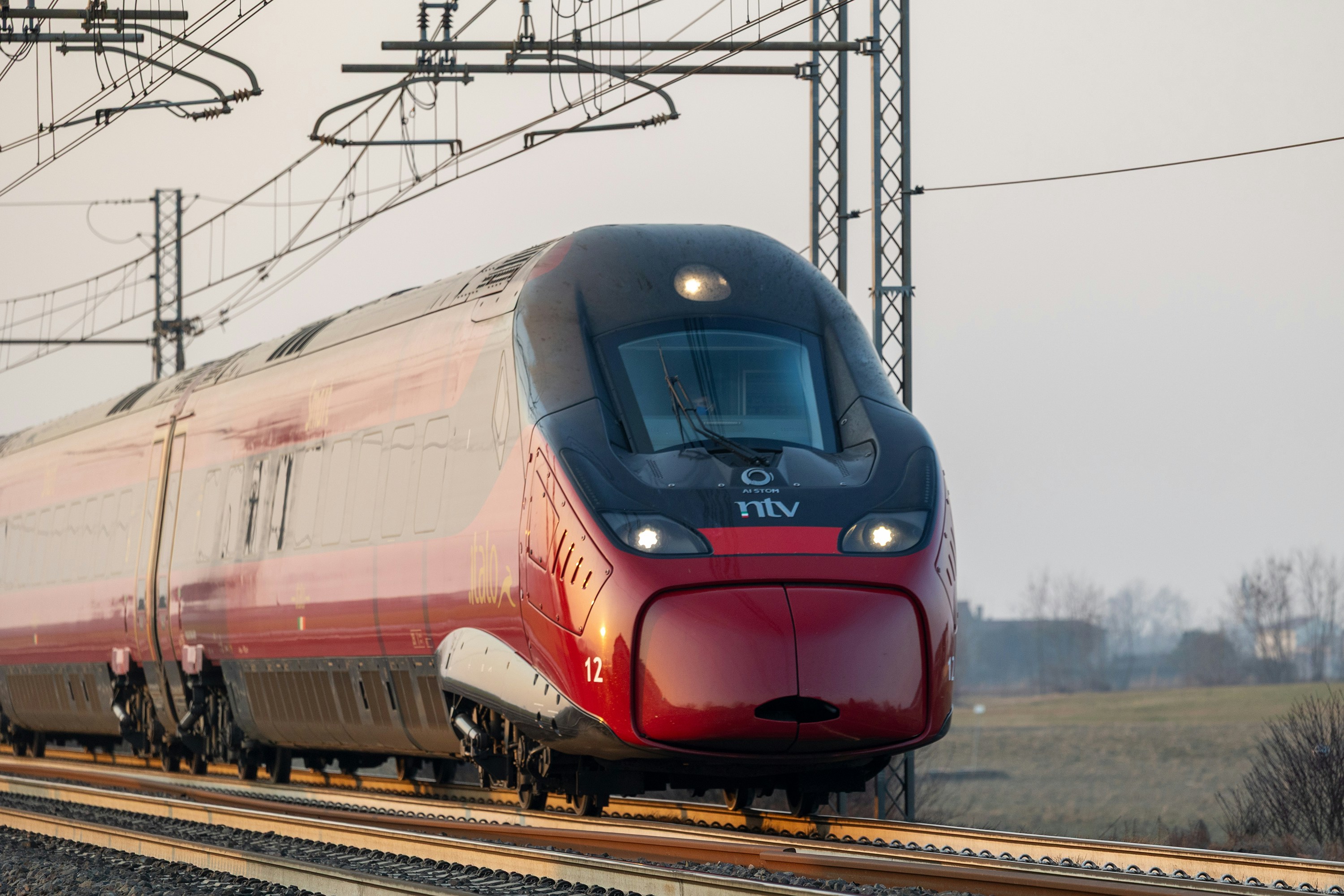 A red high-speed train traveling on tracks through a rural landscape at golden hour