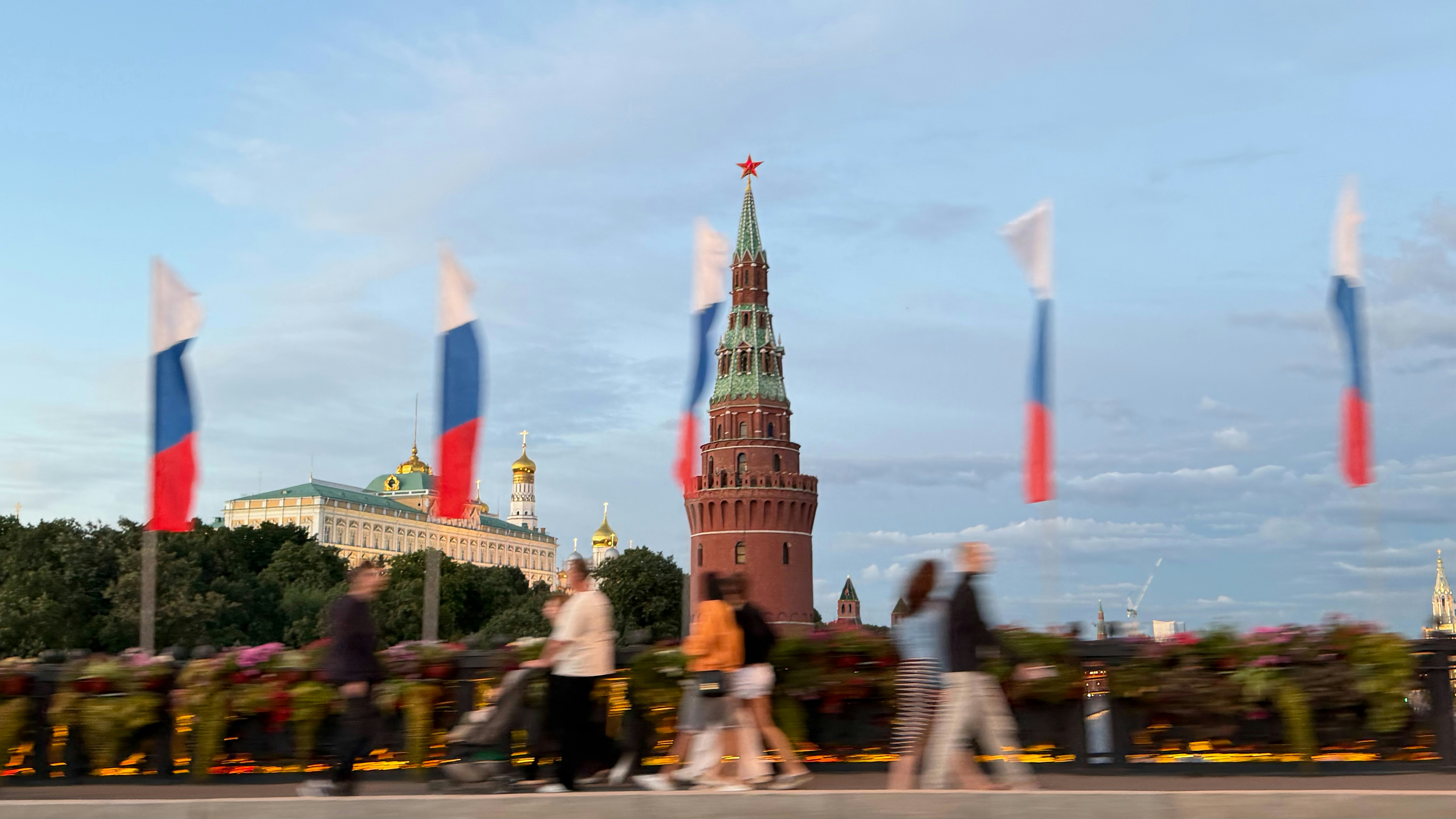 People walking past Russian flags and the Kremlin tower in Moscow at dusk