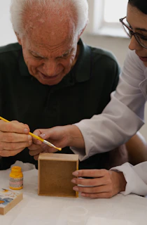An older man and a woman painting a small wooden box with yellow paint