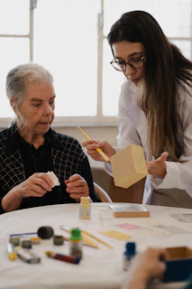 A woman in a white coat painting a wooden box for an elderly woman