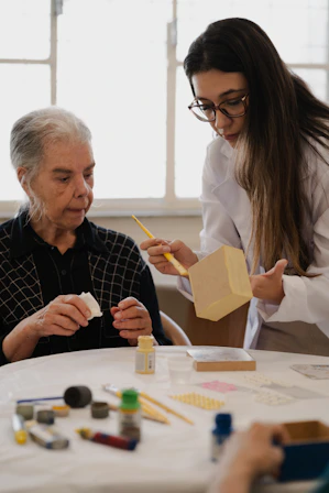 A woman in a white coat painting a wooden box for an elderly woman