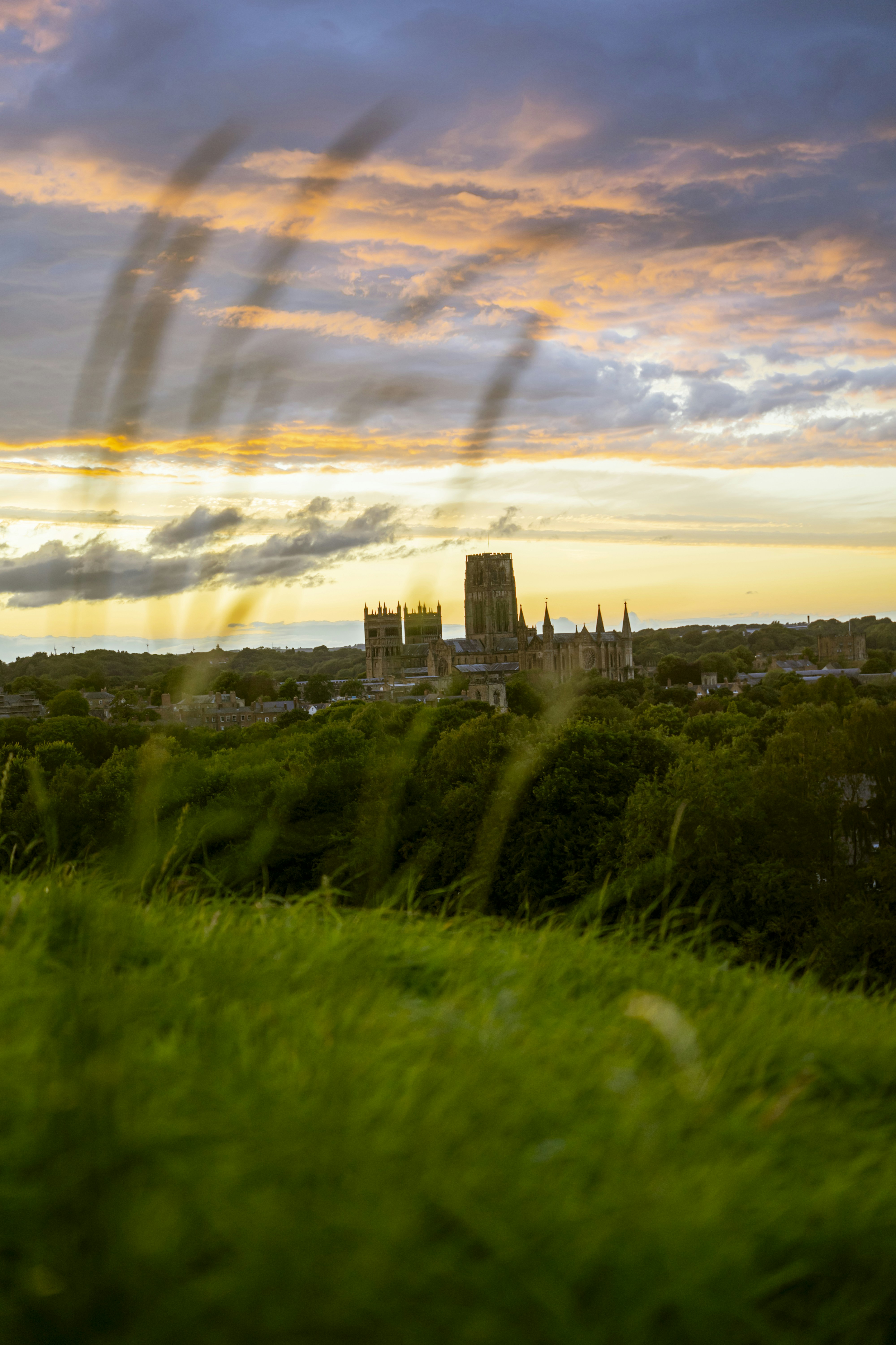 Durham Cathedral rising above green trees under a warm orange sky at golden hour