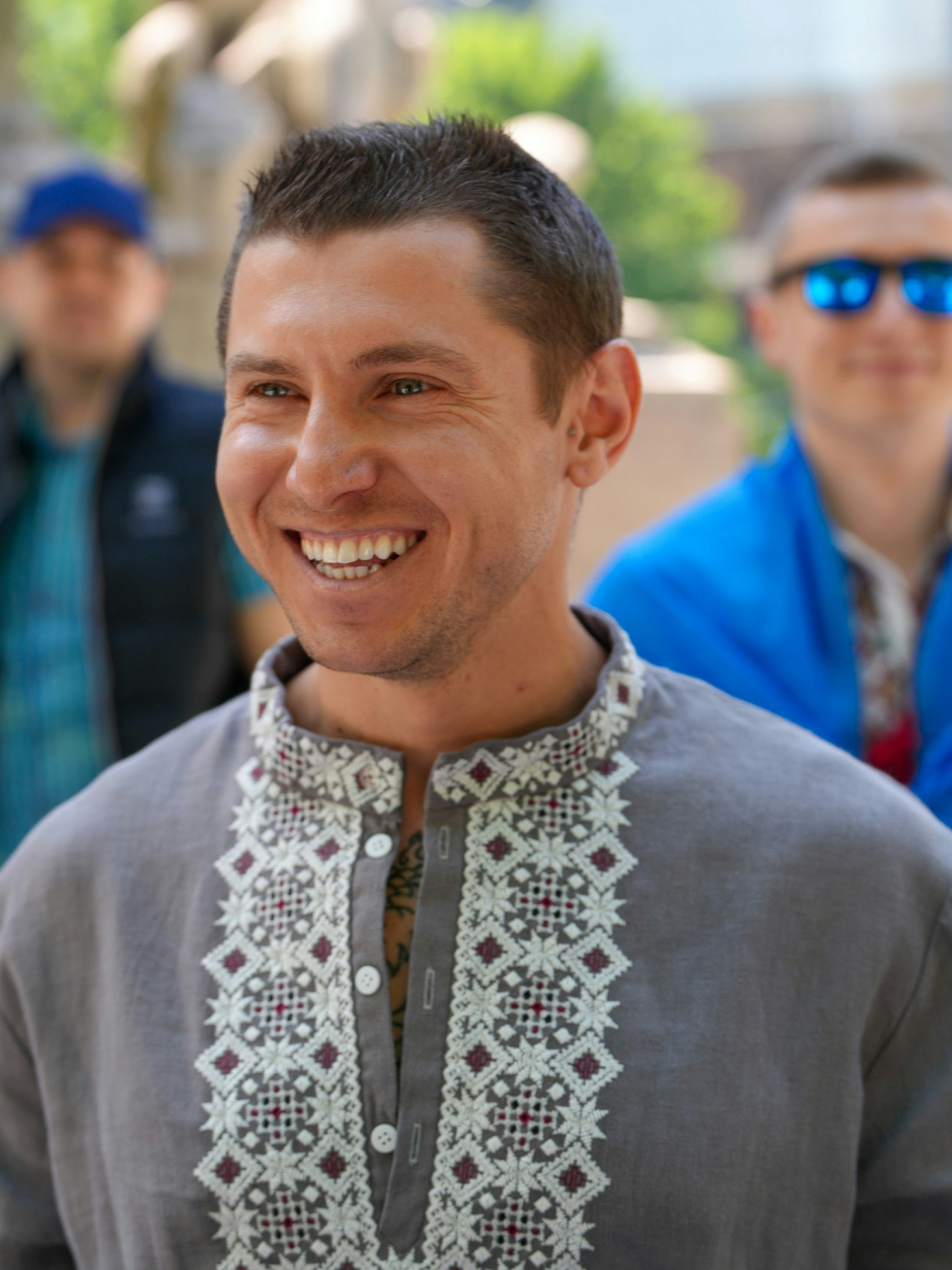 A smiling man wearing a grey embroidered shirt with two people in the background