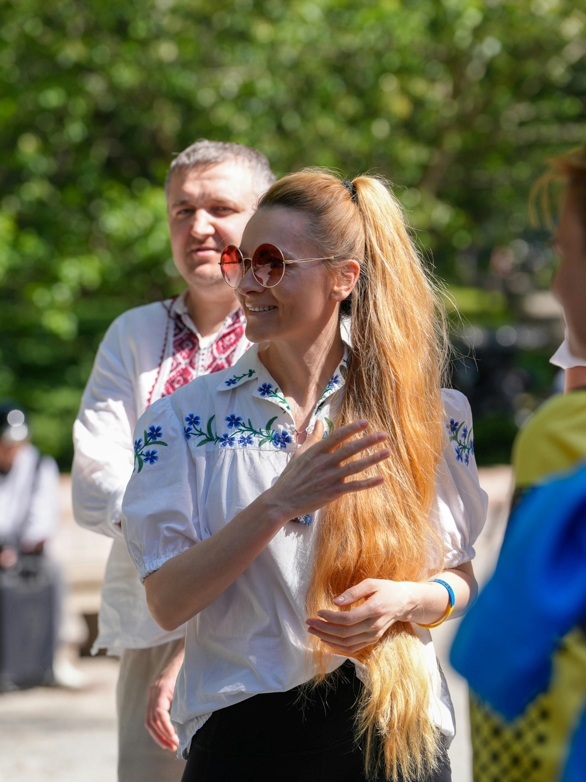 A woman with long blonde hair wearing round sunglasses and a white embroidered shirt