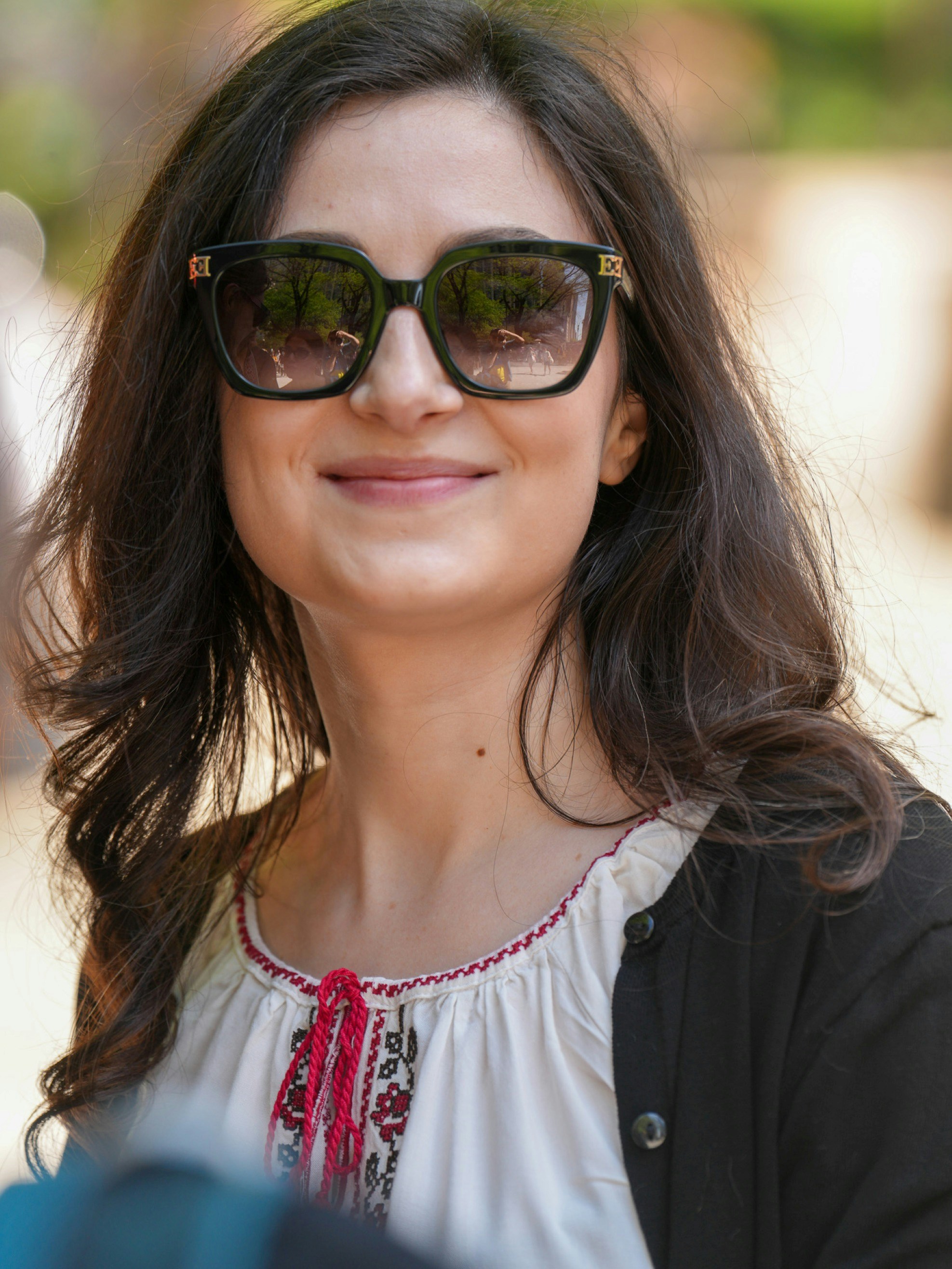 A woman with long dark hair wearing sunglasses and an embroidered white blouse