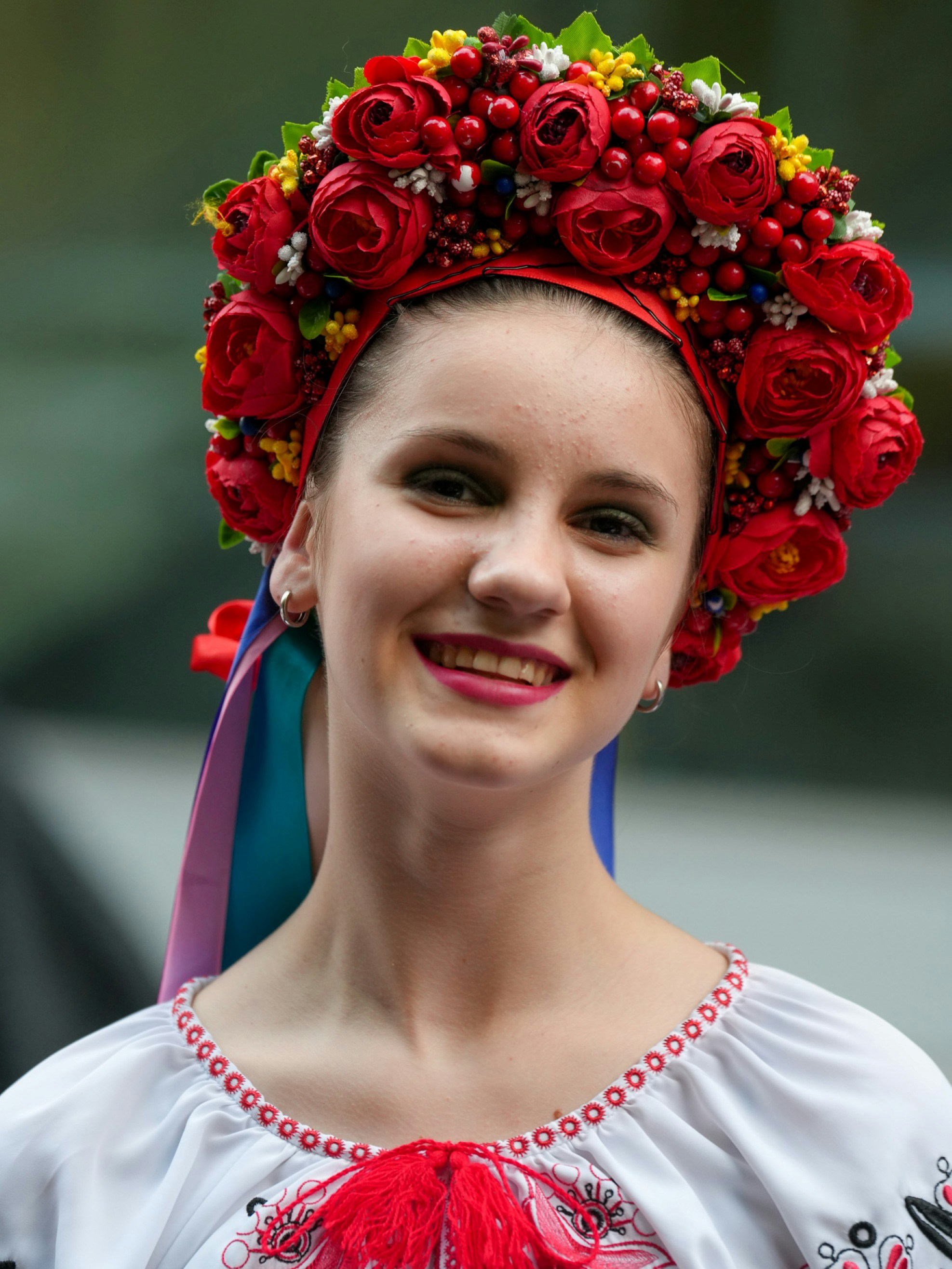 Close-up of a beaming young woman wearing a full floral crown of red roses, berries and yellow blossoms with trailing ribbons. Her white blouse features red cross-stitch trim and a red tassel, captured at a Ukrainian festival in Manhattan. Scenes from the 2025 Ukrainian Festival in Manhattan's East Village, celebrating Ukrainian culture, heritage, music, food, and community in the heart of Little Ukraine. Photo: Olek Buzunov (@buzunov.photoart)