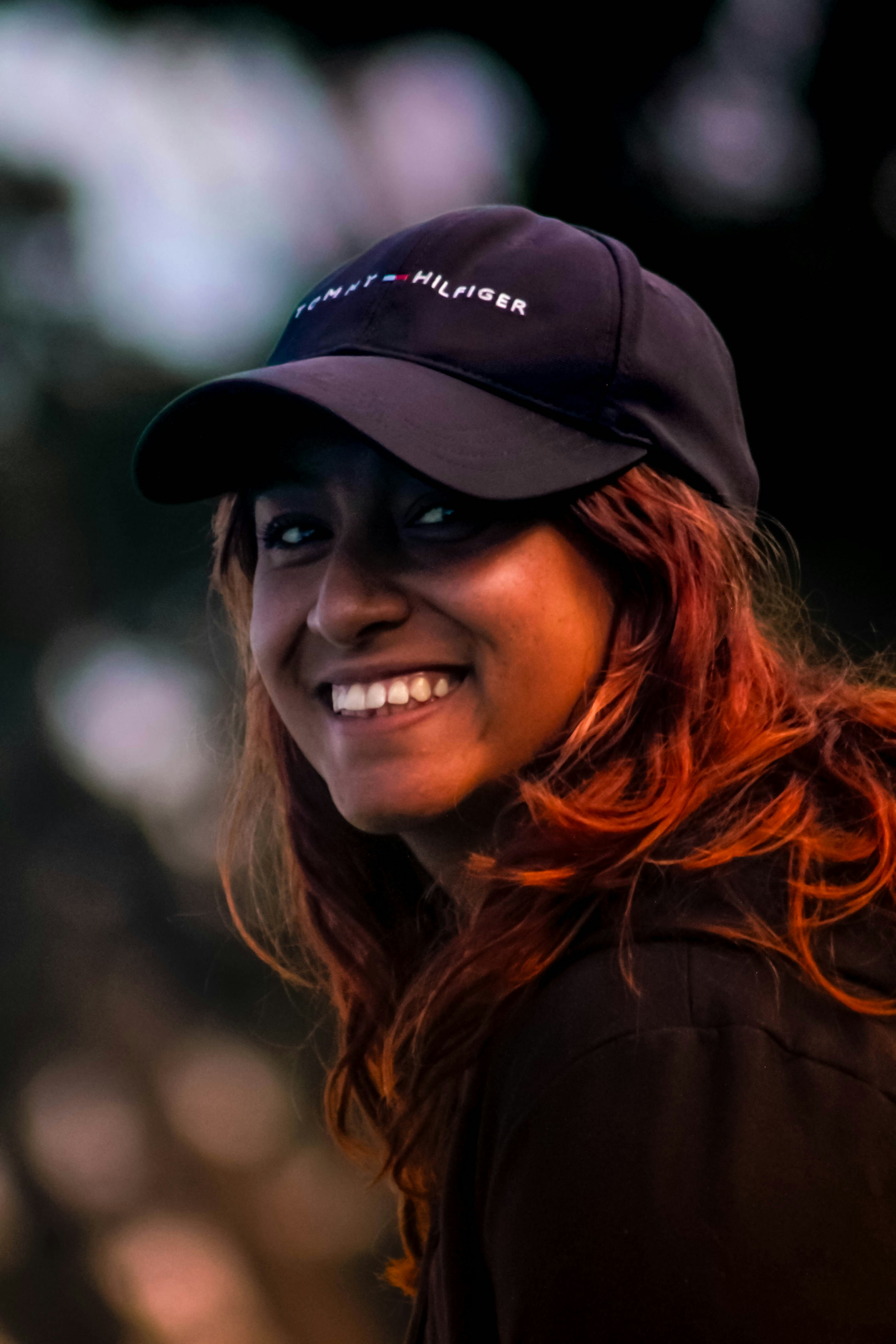 A smiling woman with reddish hair wearing a black baseball cap