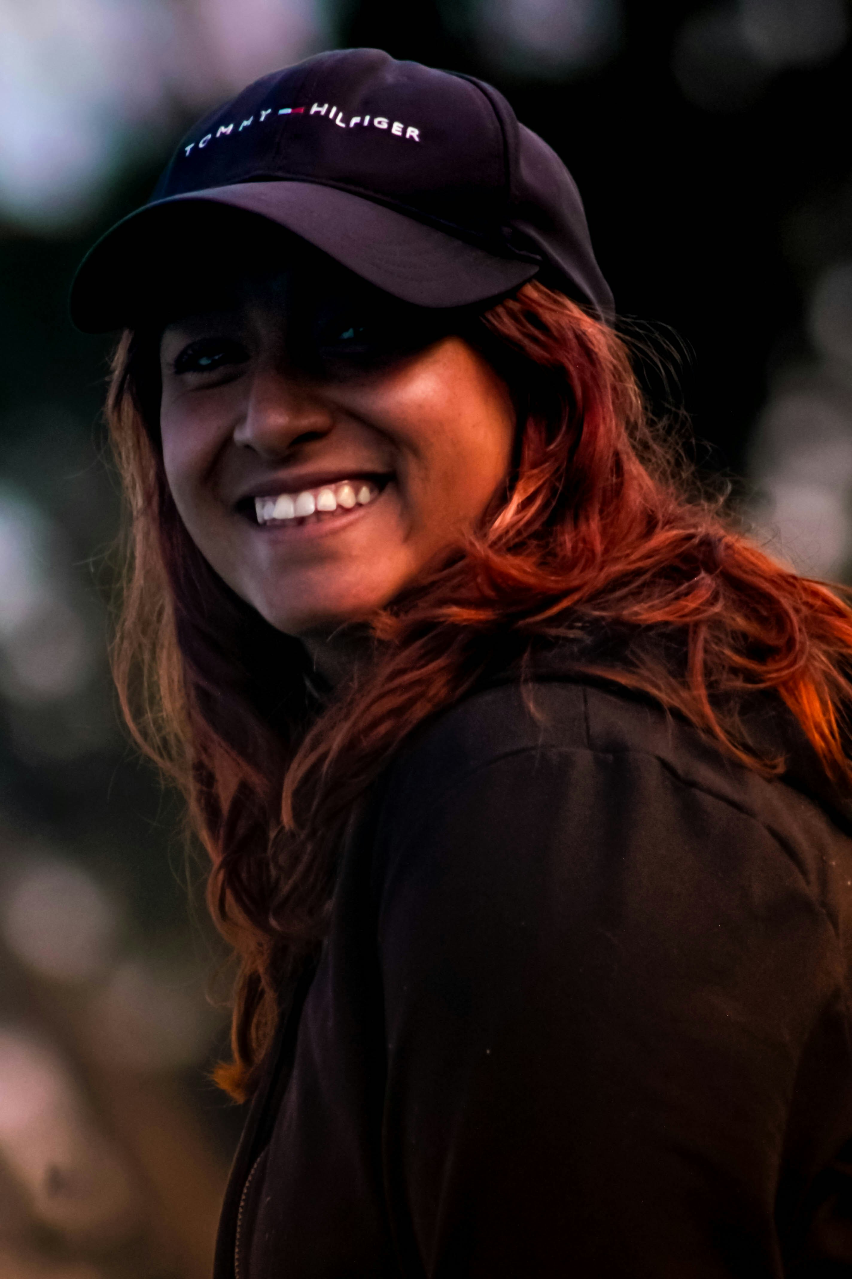 A smiling woman with reddish hair wearing a black baseball cap